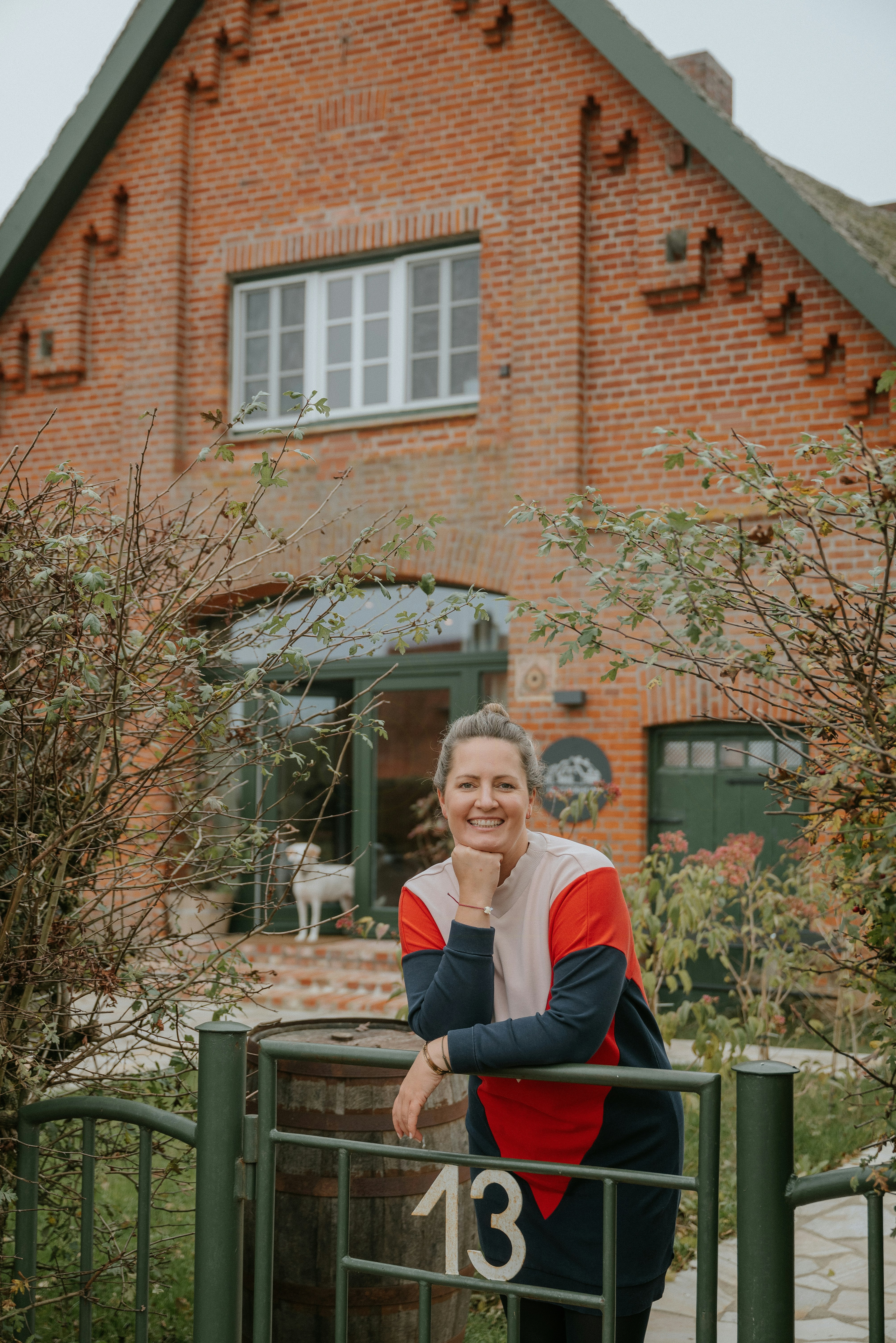 Person leaning on a green metal gate with the number 13, in front of a red brick house with green doors and windows, surrounded by bushes and plants.
