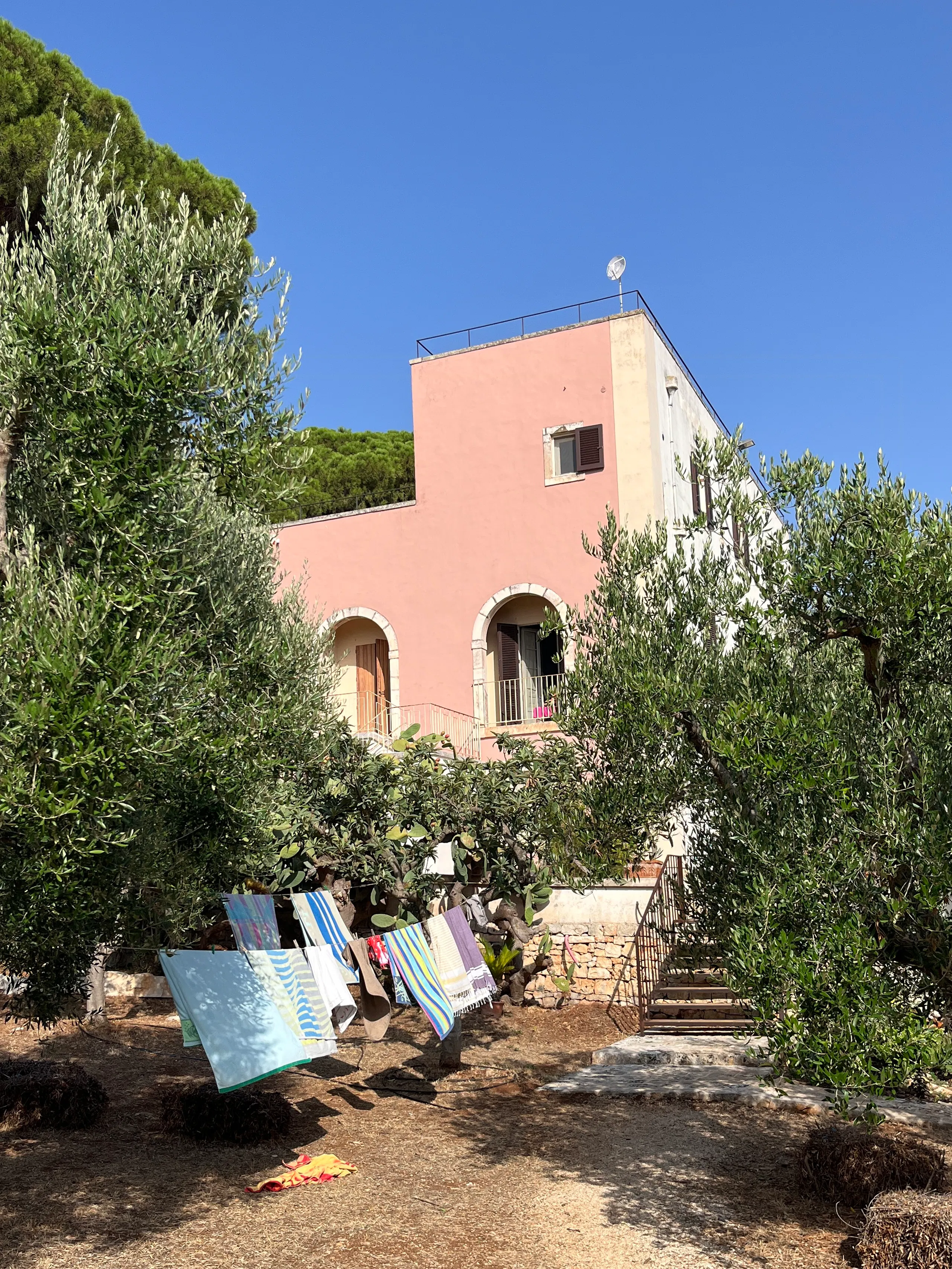 Pink stucco house with arched balcony openings, surrounded by olive trees and greenery, with colorful towels hanging on a clothesline in the foreground