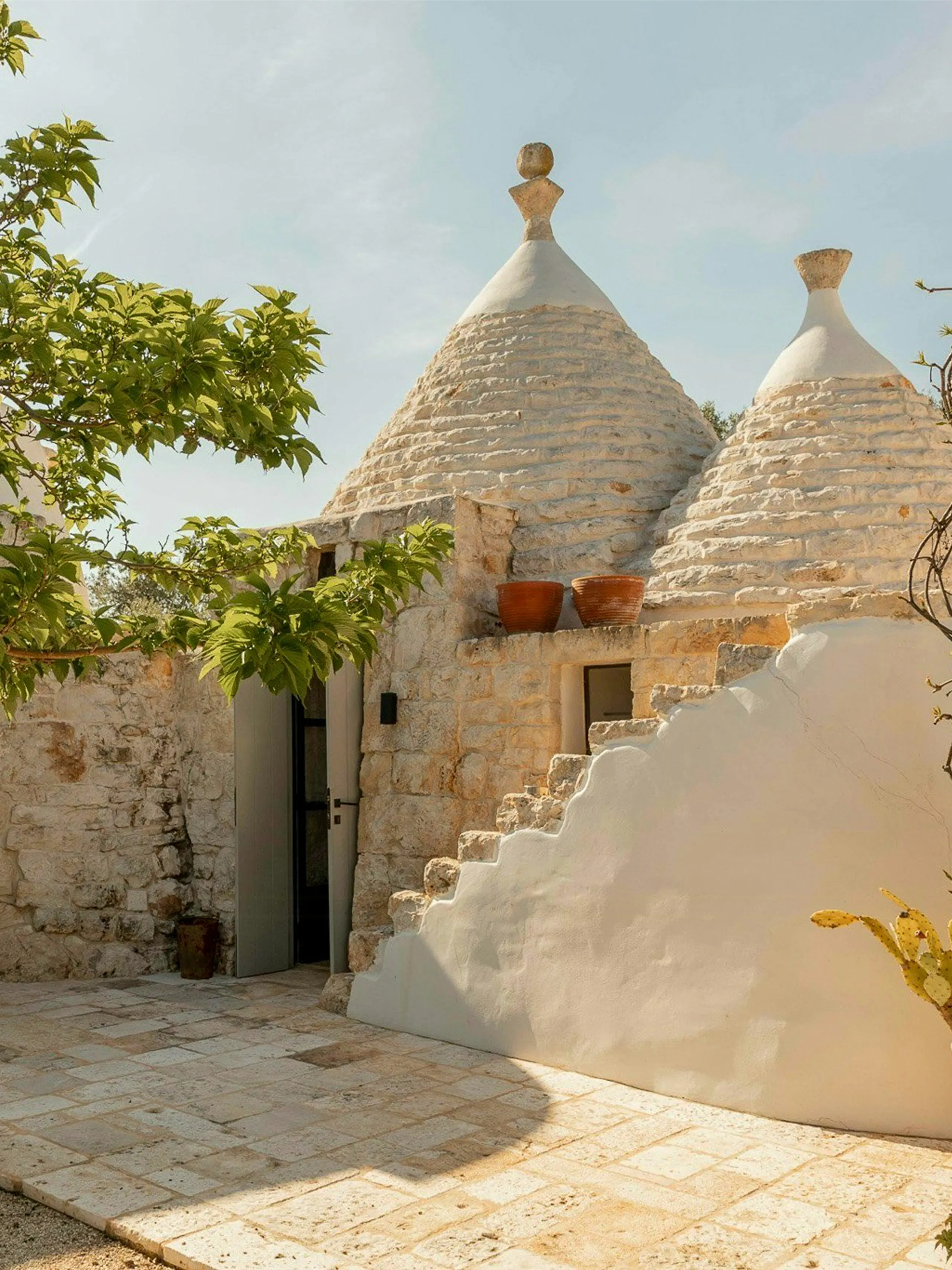 Stone trullo-style house with two conical roofs, open doorway, terracotta pots on a ledge, white stucco stair wall, and tiled courtyard with leafy tree branches nearby