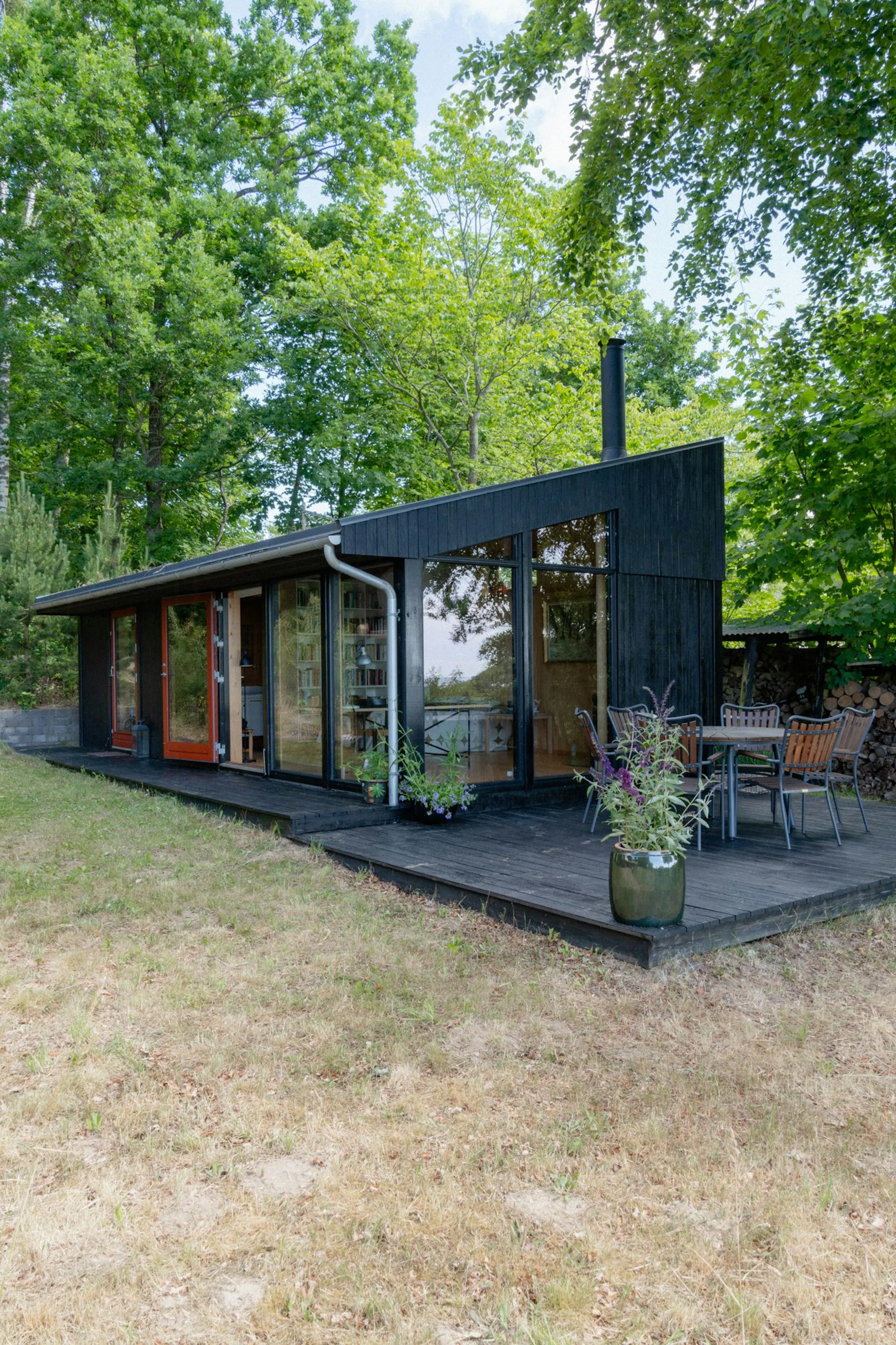 Black modern cabin with large windows, slanted roof and wooden deck with outdoor dining table, surrounded by trees and grass