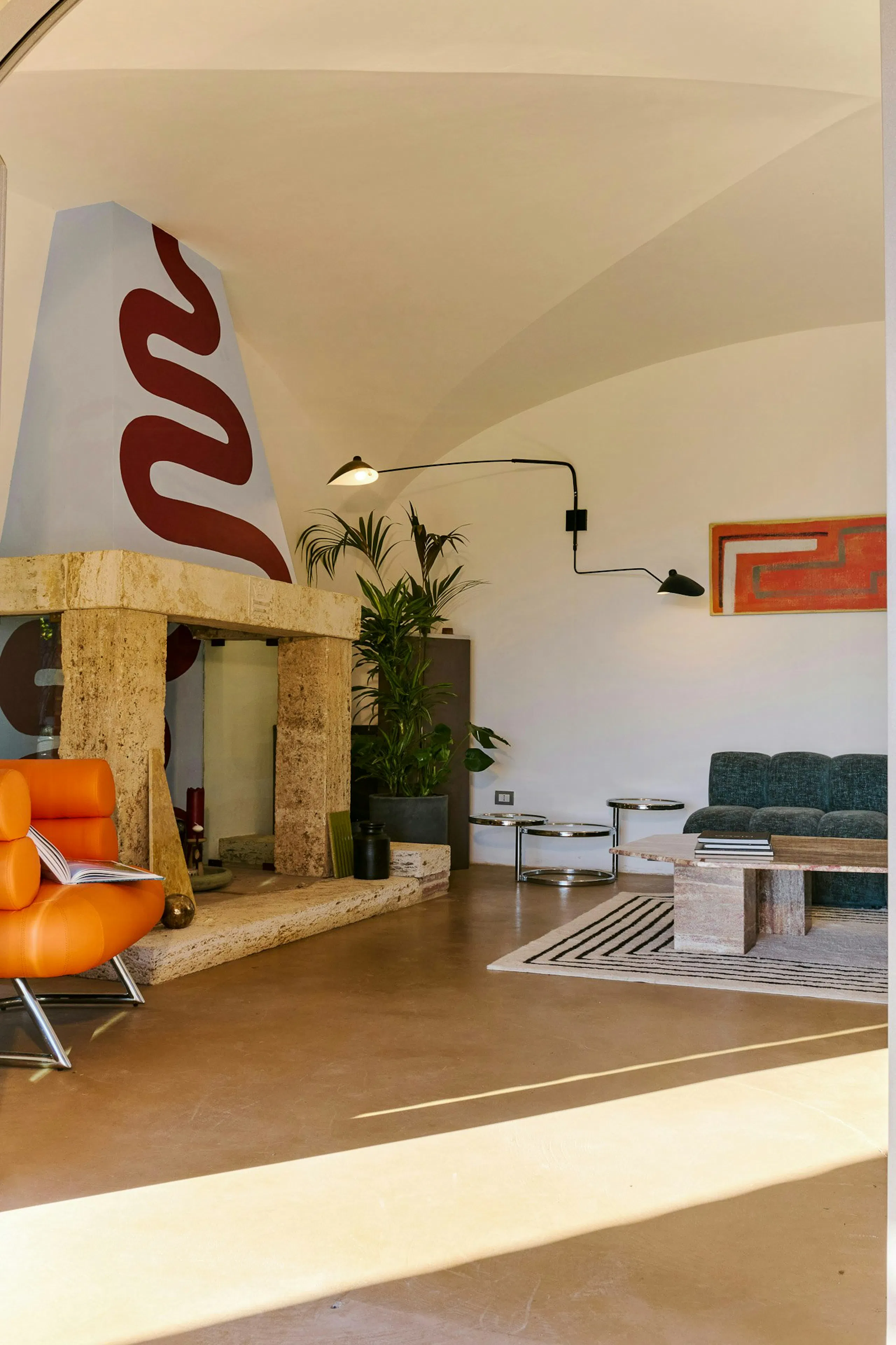 Modern living room with a sculptural stone fireplace, orange lounge chair, teal sofa, wall-mounted lamps, potted plants, coffee tables, and a curved white ceiling