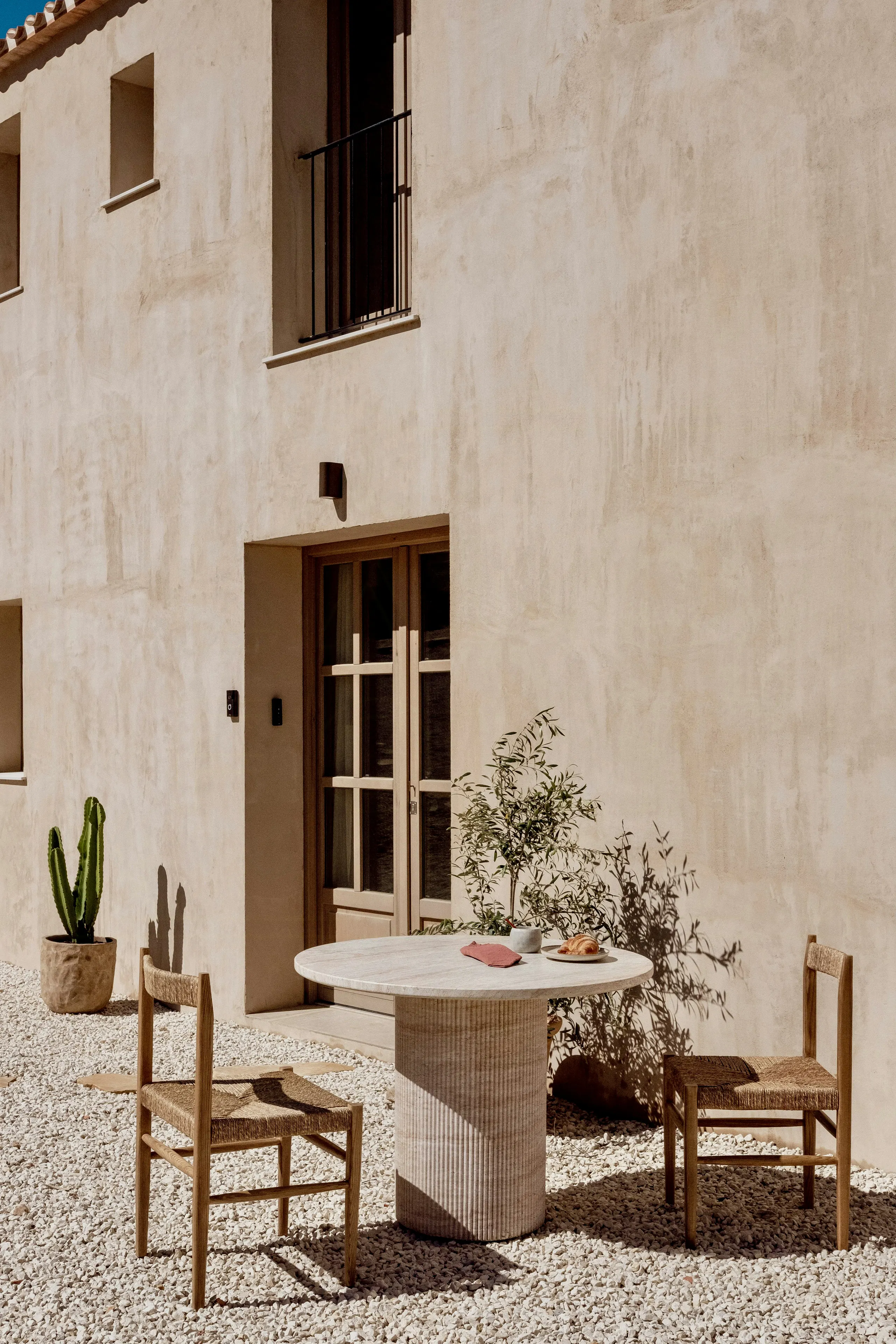 Minimal outdoor patio with beige stucco wall, French doors, round stone table, two wooden chairs, potted cactus, and gravel ground cover