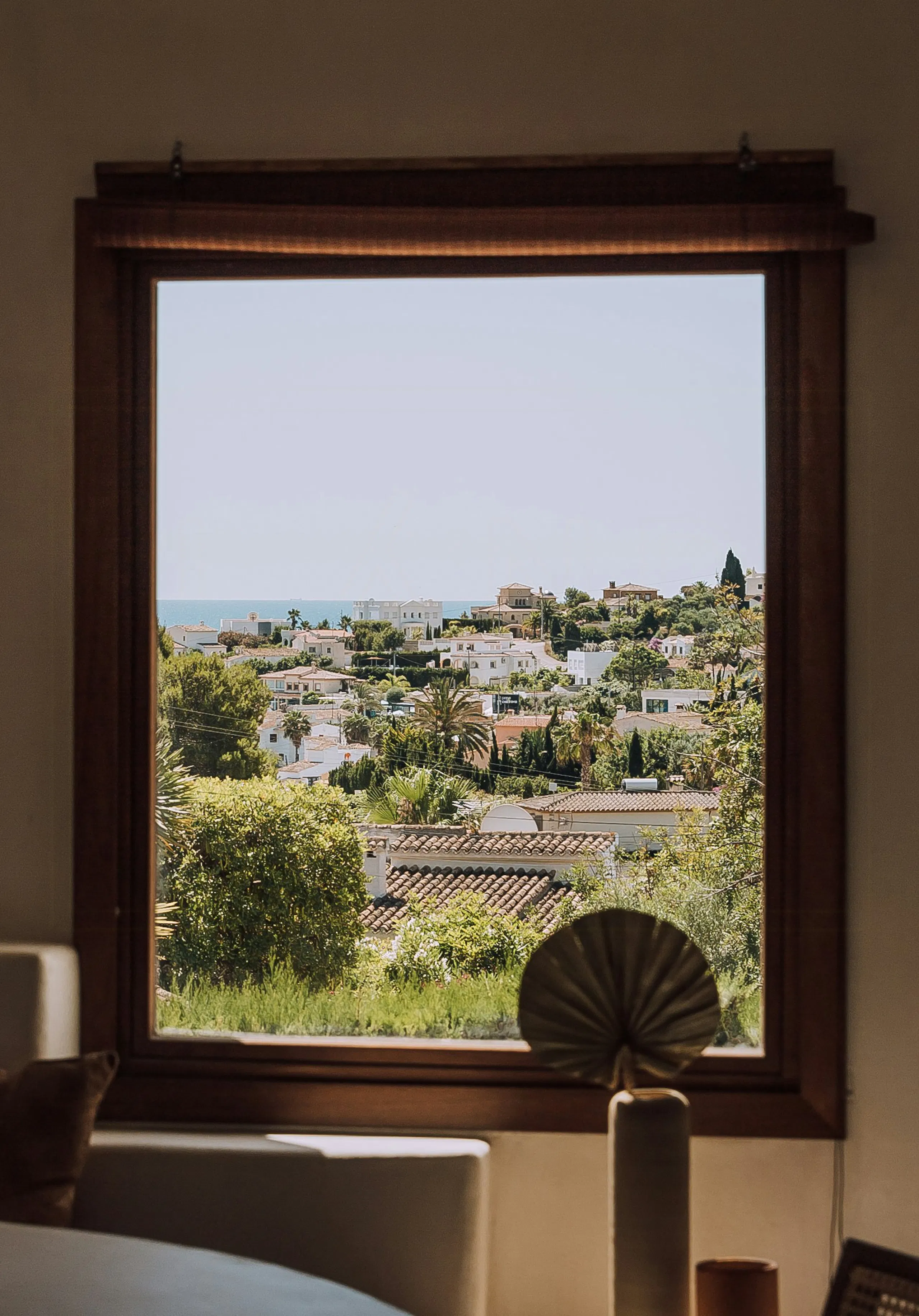 View through a wooden-framed window of a hillside neighborhood with houses, trees, and the sea in the distance, with indoor furniture in the foreground