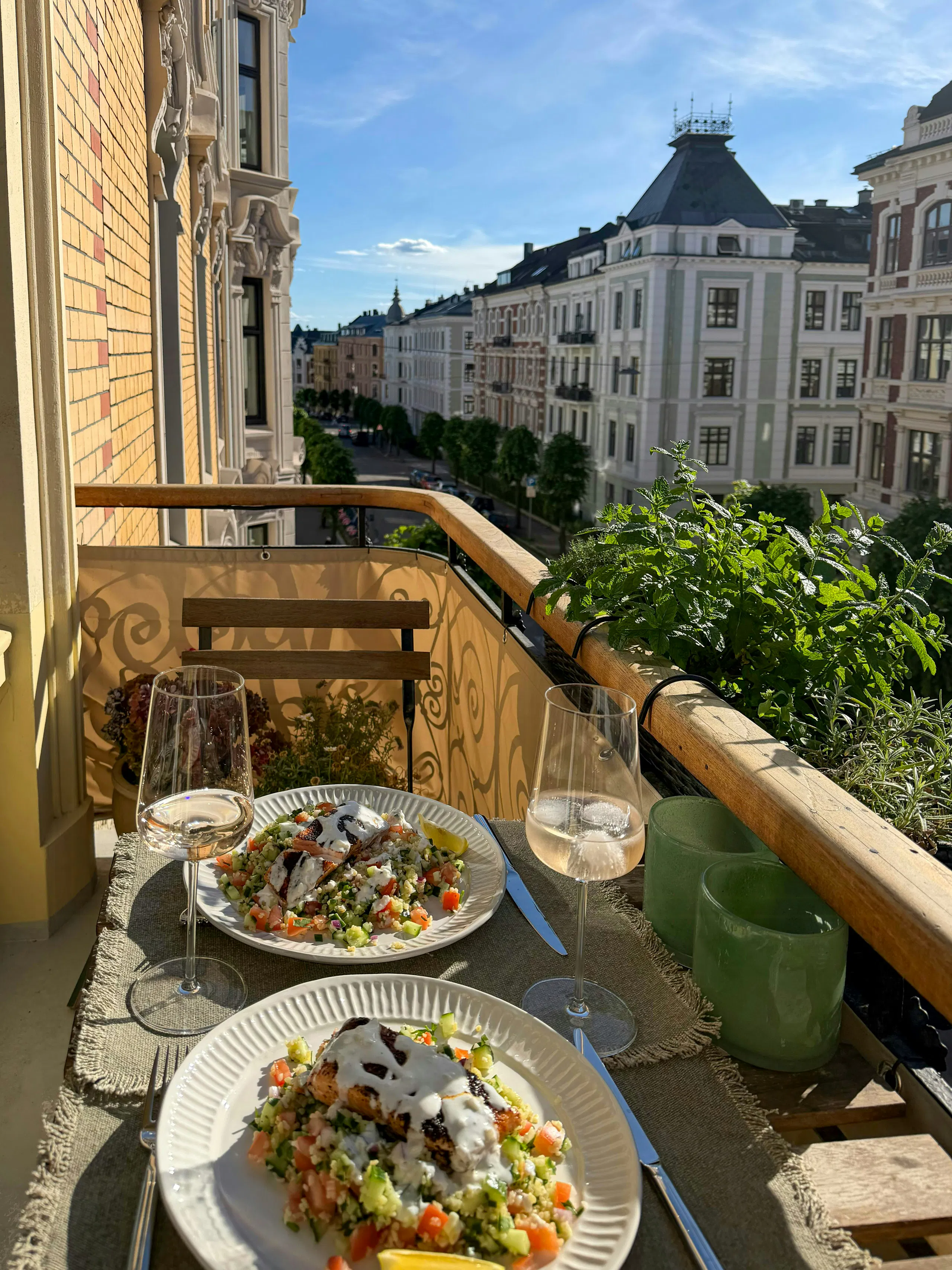 Balcony table set with two plates of salad, wine glasses, and green candles overlooking a city street lined with historic apartment buildings