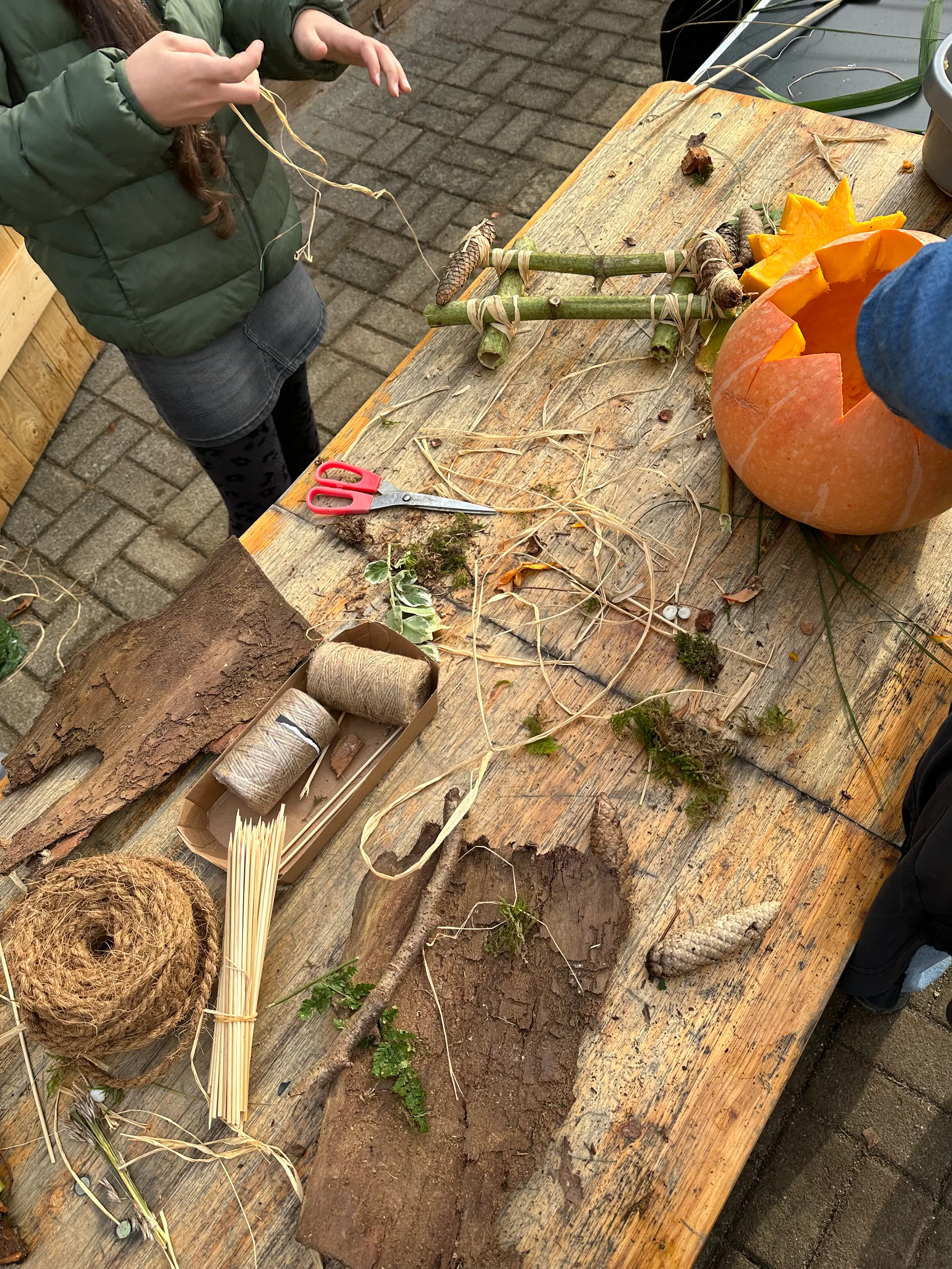 Children crafting with natural materials on a weathered wooden table, with scissors, twine, sticks, straw, moss, and a carved pumpkin on a paved patio