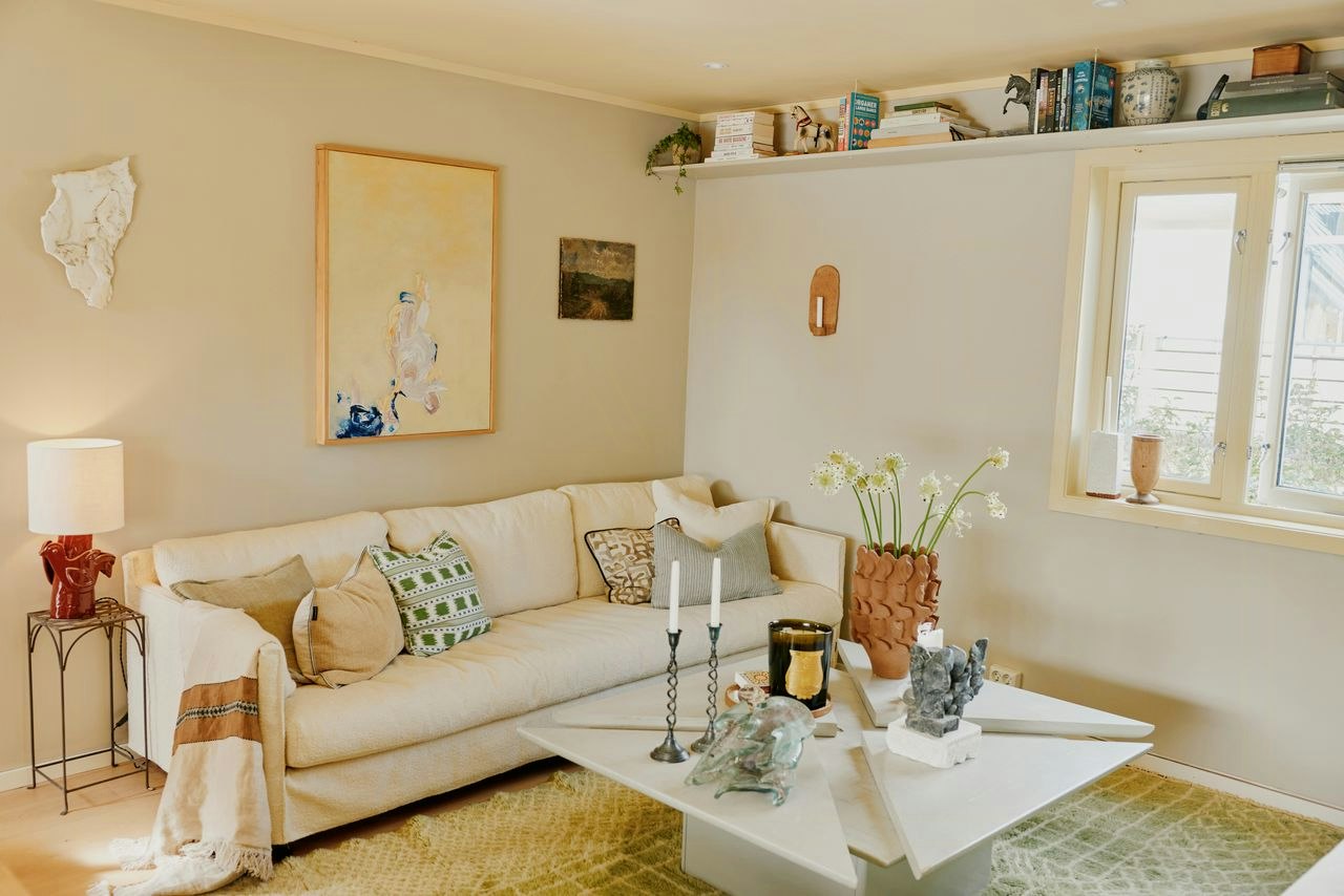 Living room with a cream sofa, abstract wall art, floor lamp, white geometric coffee table, window, wall shelf with books, and patterned rug