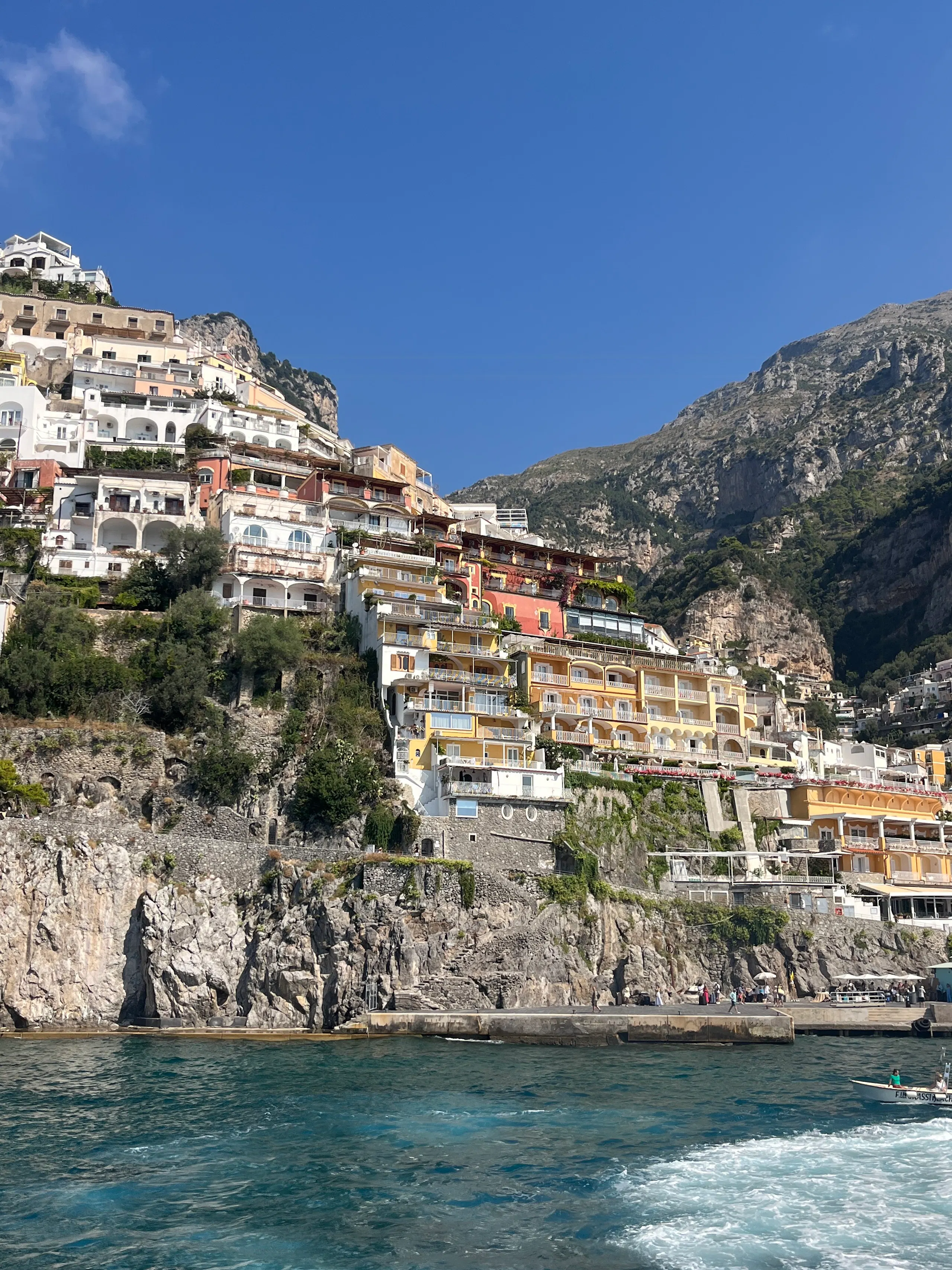 Colorful terraced buildings built into a steep rocky hillside above a blue sea, with mountains and a clear sky in the background