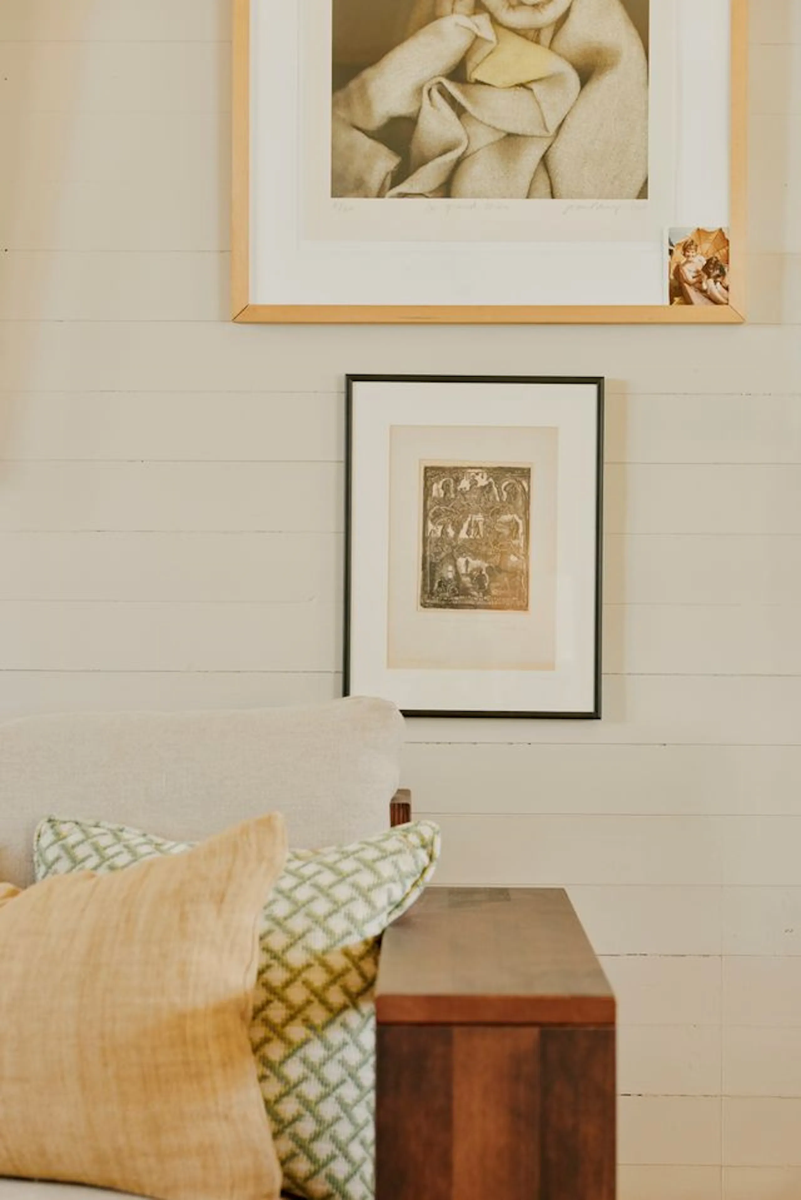 Living room corner with cream shiplap wall, two framed artworks, beige sofa, patterned cushions, and a wooden side table