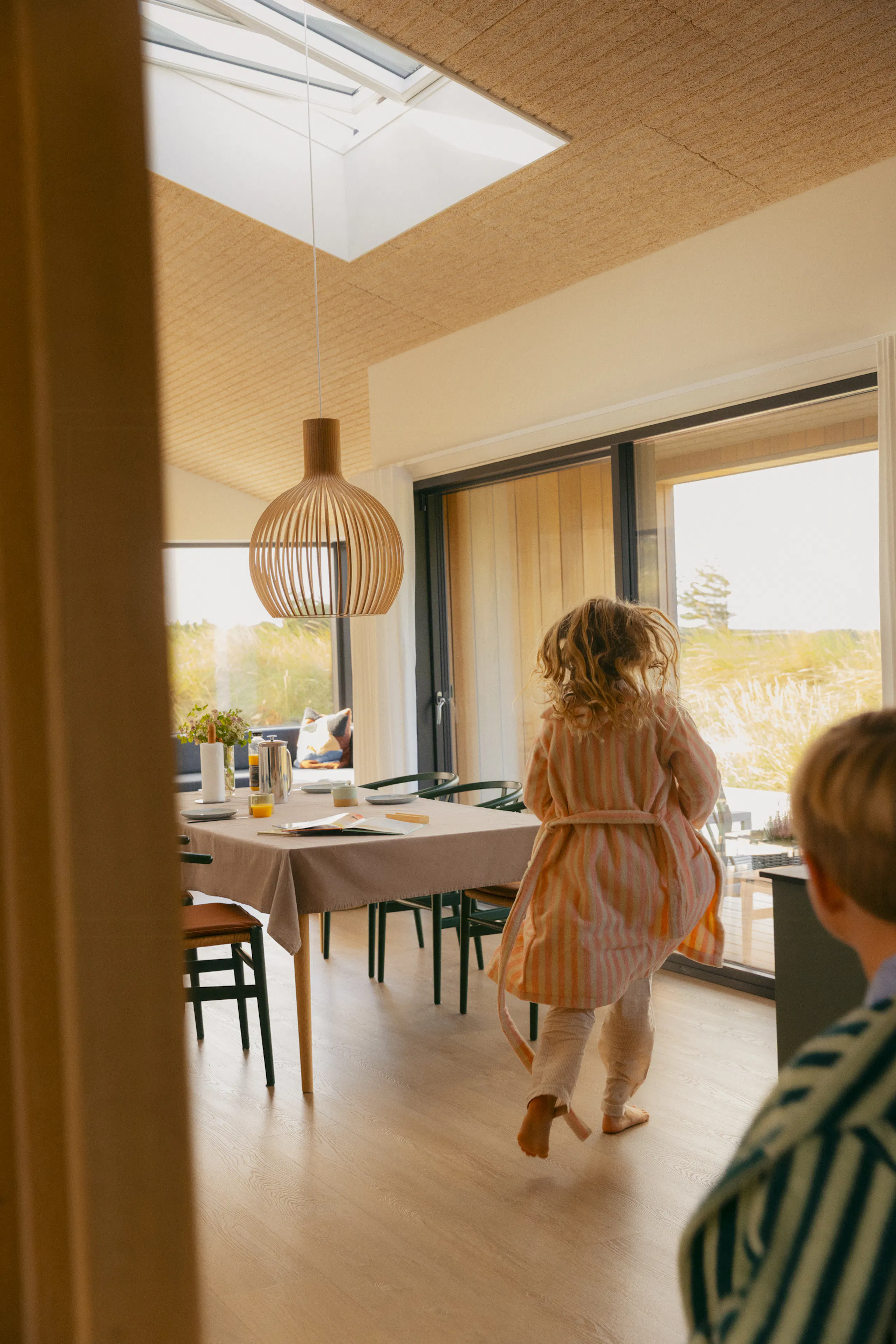 Helles Esszimmer mit Dachfenster, geflochtener Pendelleuchte, großen Glasschiebetüren und Kindern, die neben einem gedeckten Tisch mit grünen Stühlen spielen