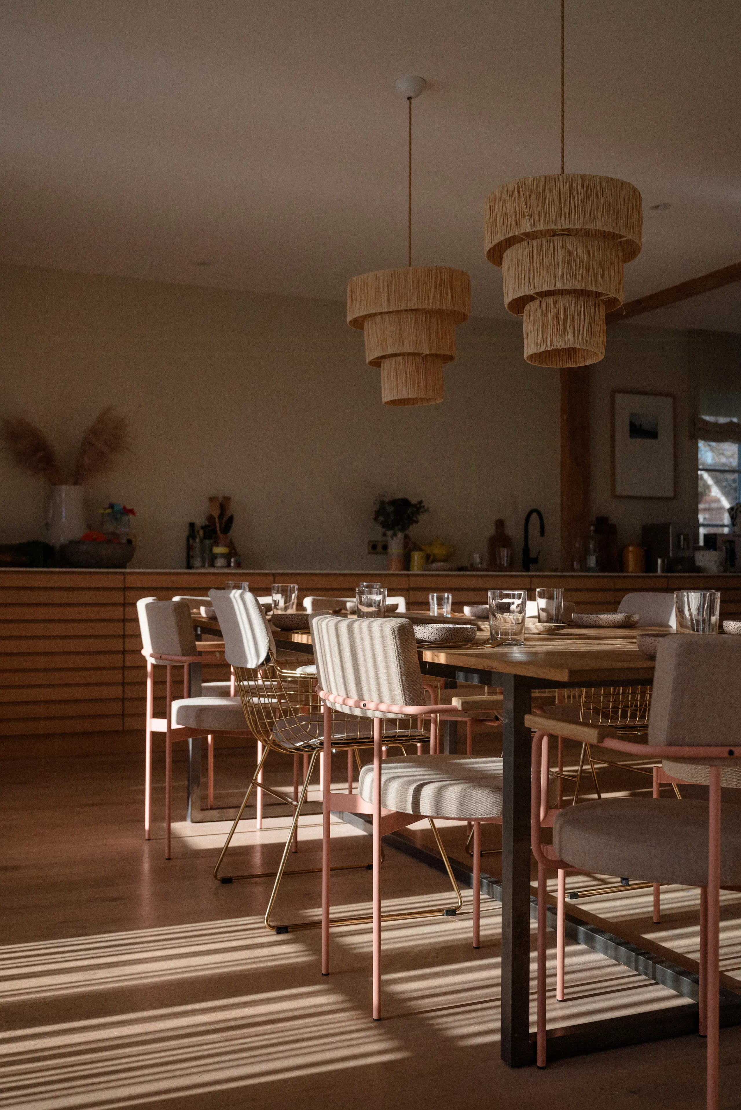 Dining area with wooden table, cushioned chairs with pink and gold frames, two layered rattan pendant lights, and a modern kitchen in the background with wooden cabinetry and decorative vases.