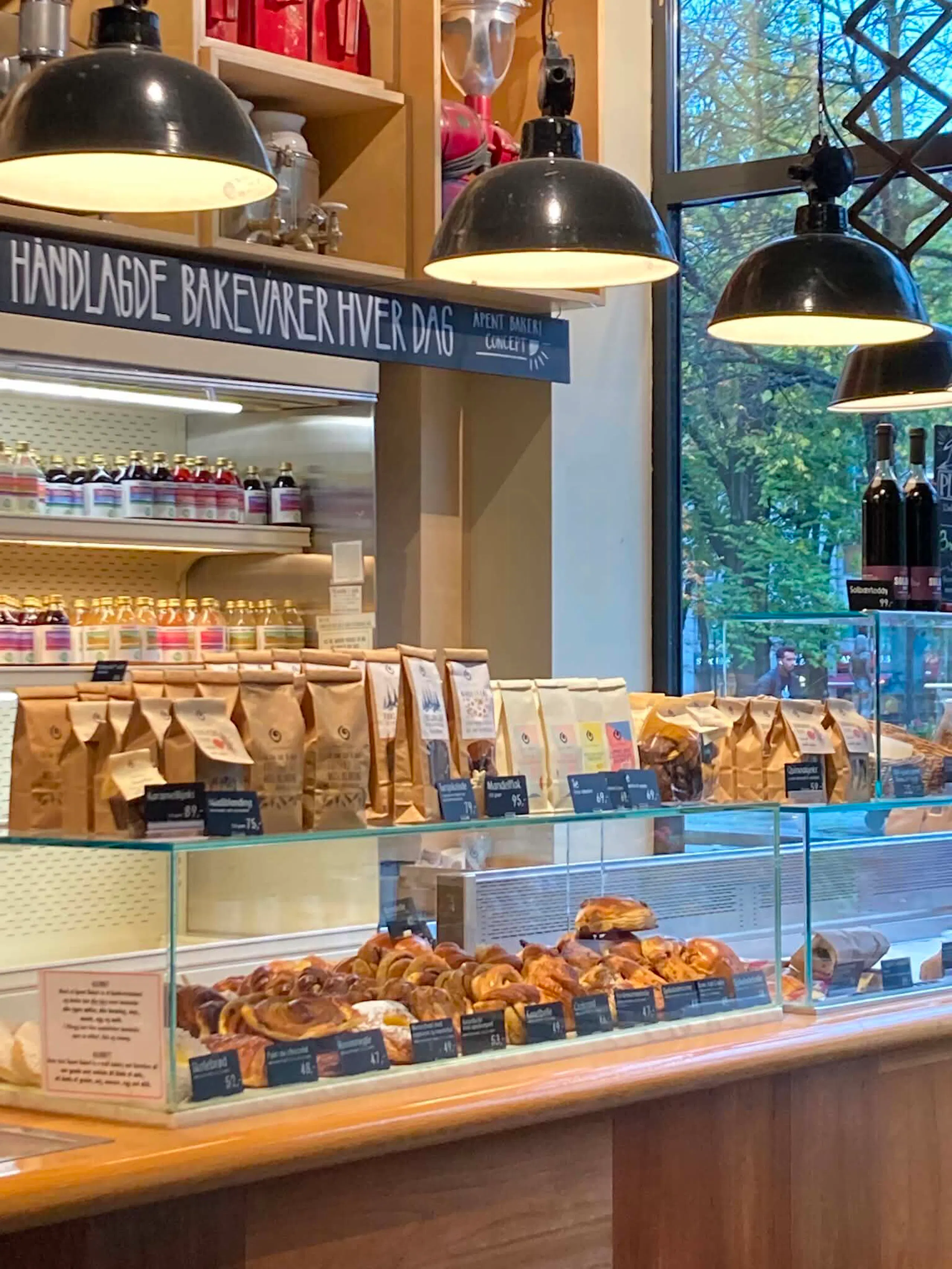 Bakery counter with glass display case of pastries and bread bags, hanging black lamps, and large window overlooking trees outside