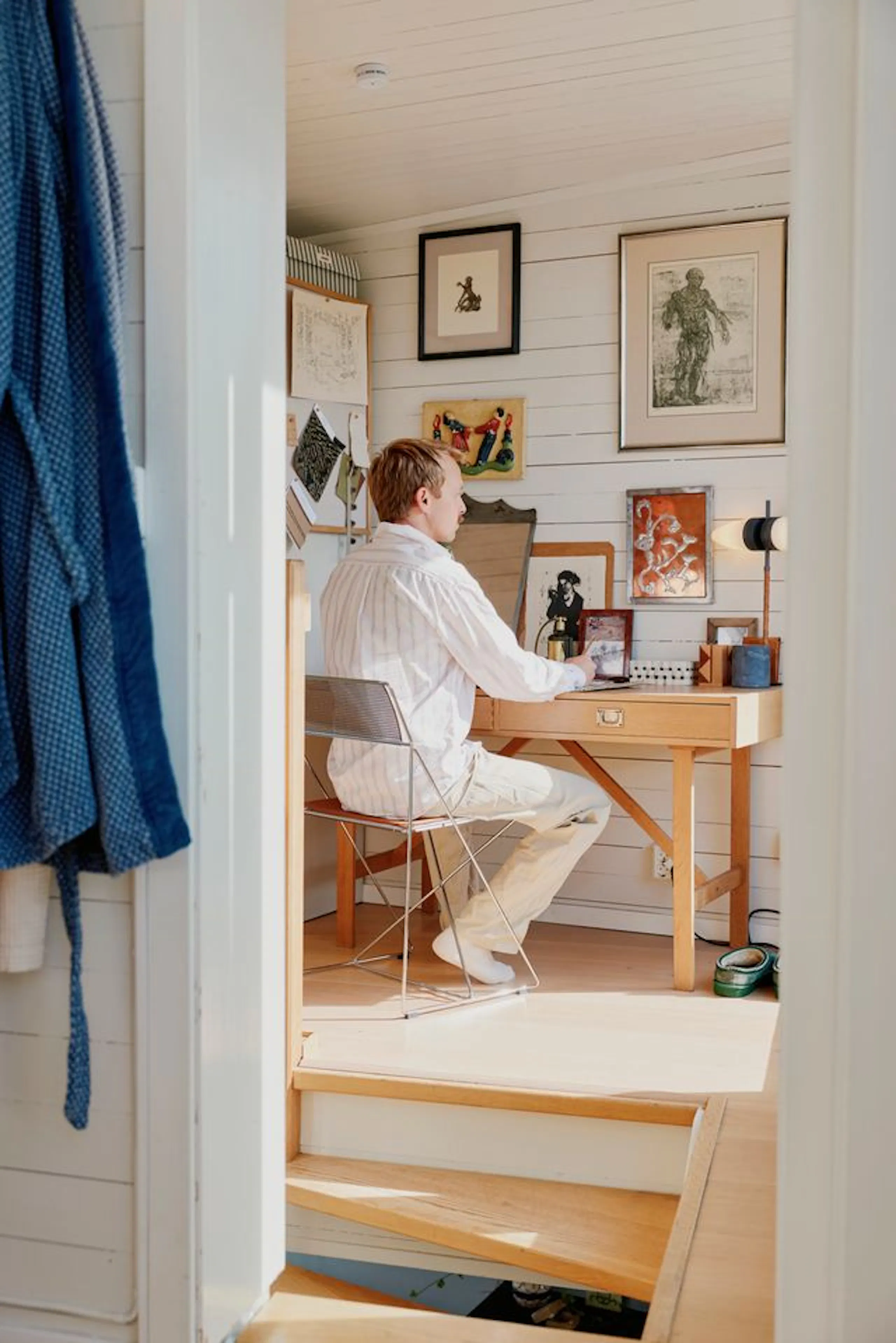 Person seated at a wooden desk in a small white-paneled home office, with framed artwork on the wall, a wire chair, and sunlight on the wooden floor