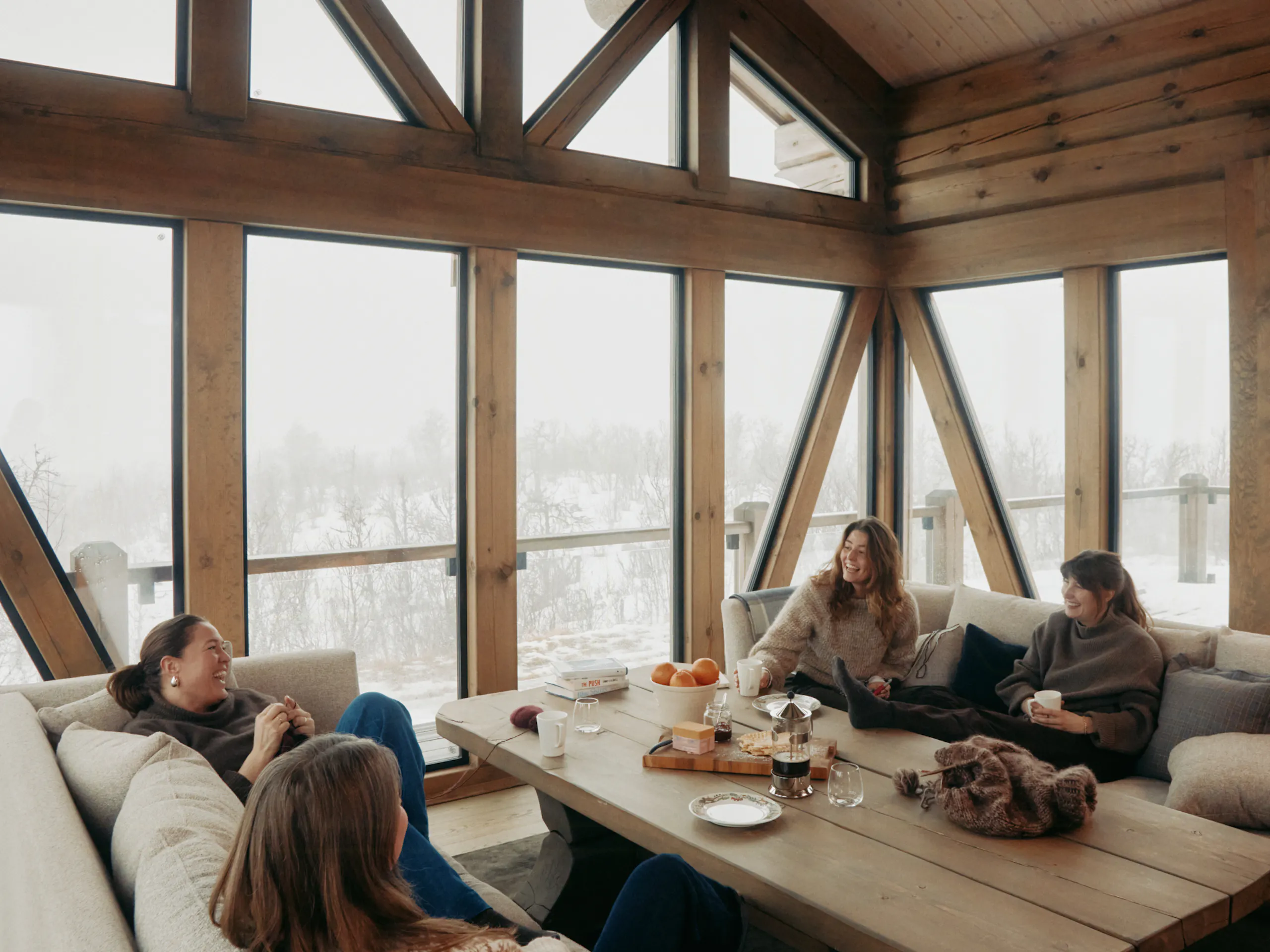 Wood-paneled living room with large floor-to-ceiling windows, triangular beams, four people sitting on beige sofas around a wooden coffee table, with a snowy landscape outside