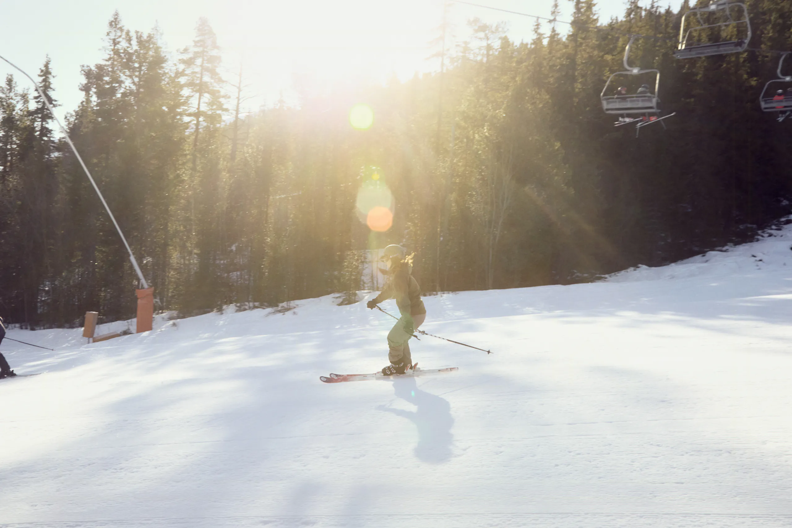 Skier descending a snowy slope in bright sunlight with forested trees and ski lift chairs on the hillside