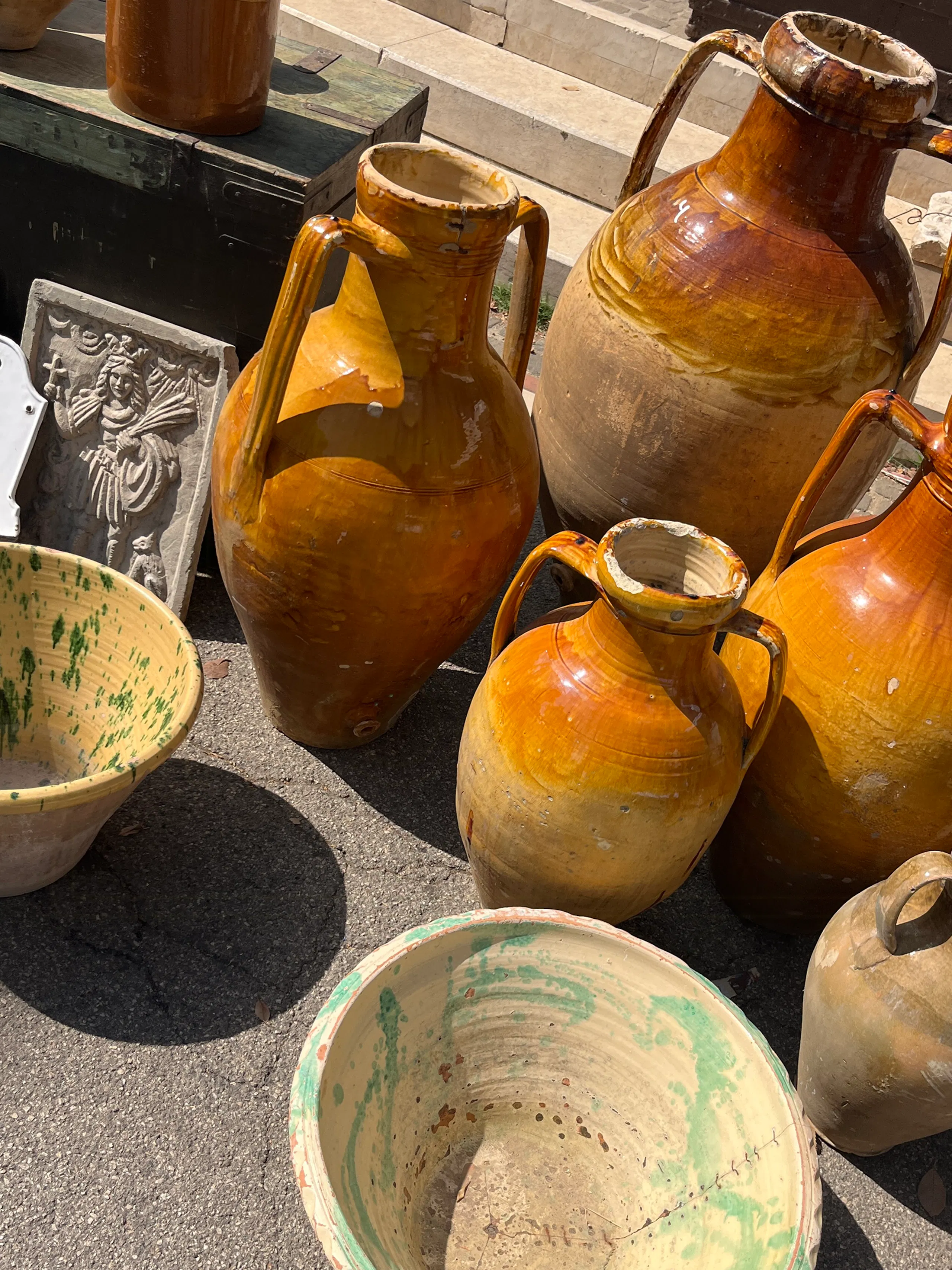 Several large amber-glazed ceramic jugs and painted bowls arranged on pavement beside a small carved stone relief and a dark crate