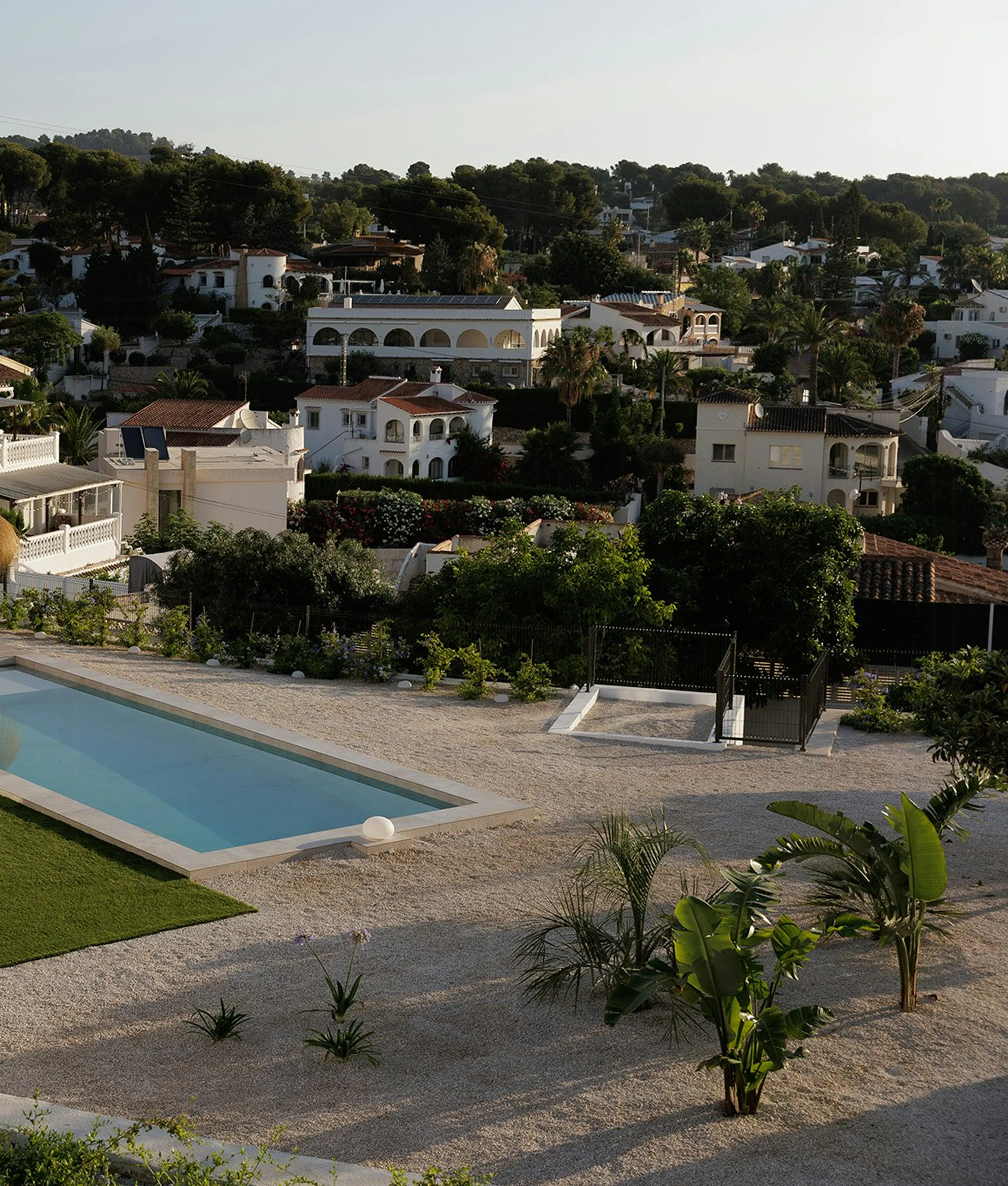 swimming pool beside a gravel terrace with small palm plants, overlooking a hillside neighborhood of white houses and trees