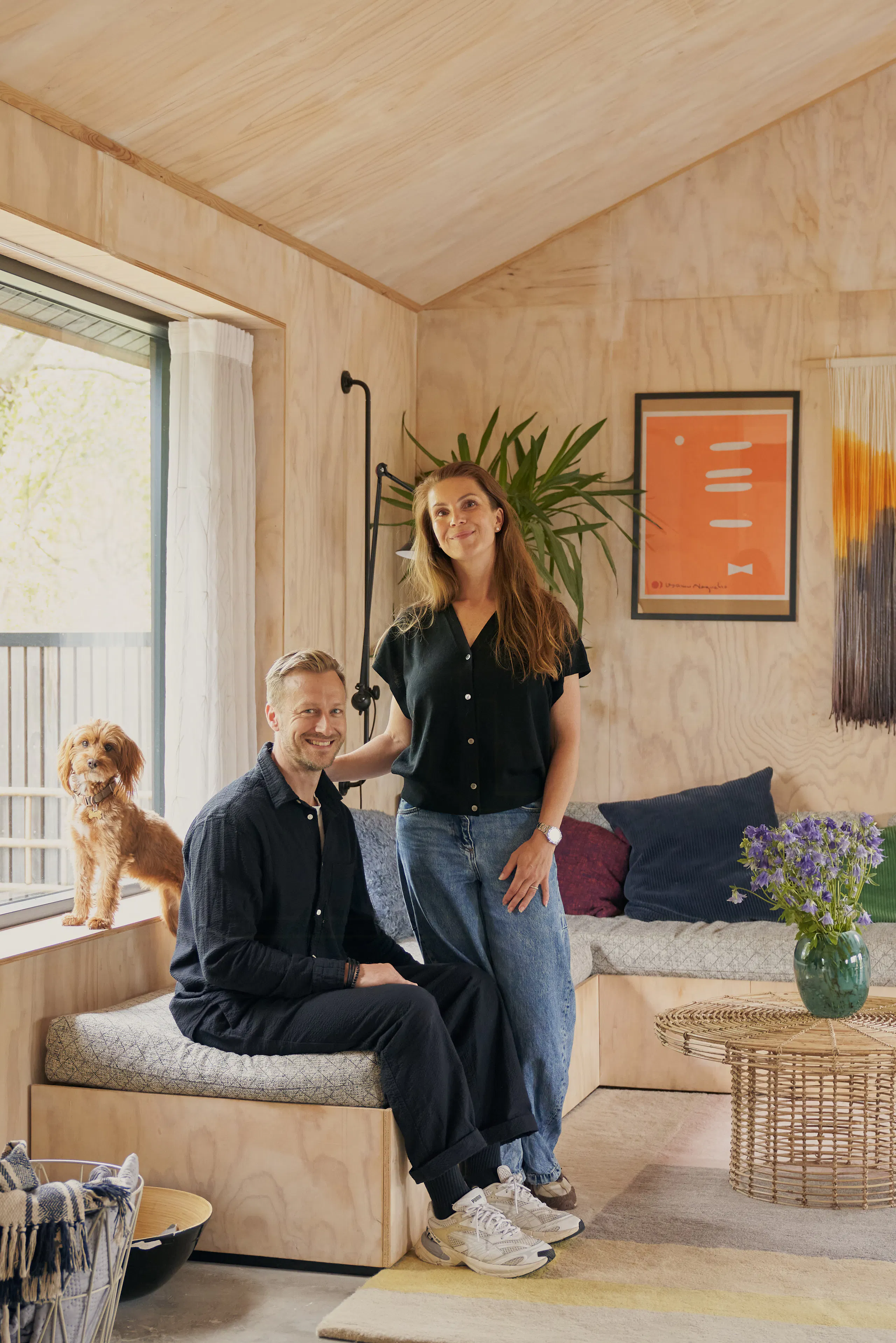 Træbeklædt stue med et par og en lille hund ved en vinduesplads, sofa med puder, kunst på væggen, grøn plante og flettet sofabord