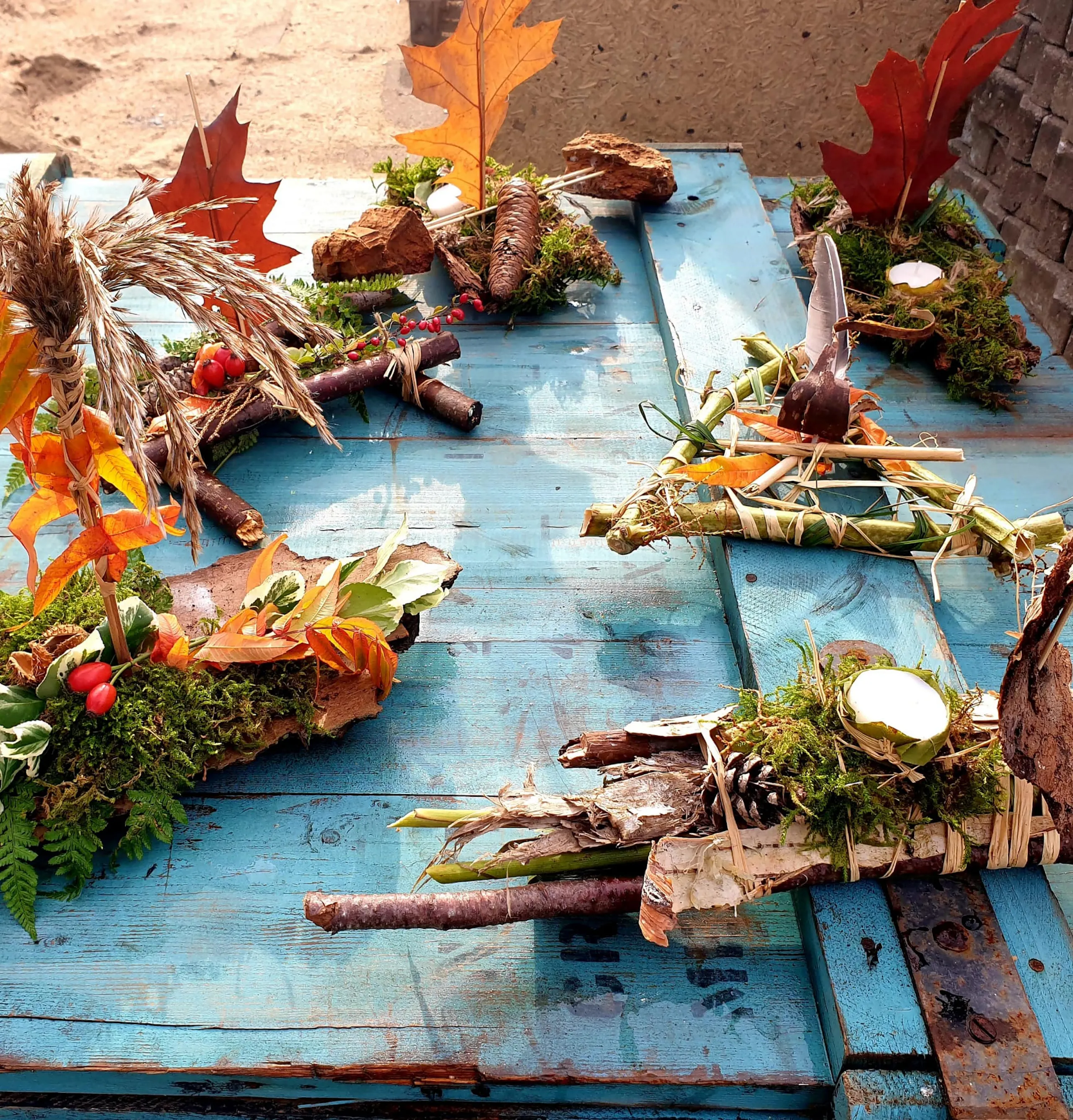 Handmade autumn decorations with sticks, moss, leaves, berries, and bark arranged on a weathered blue wooden table outdoors