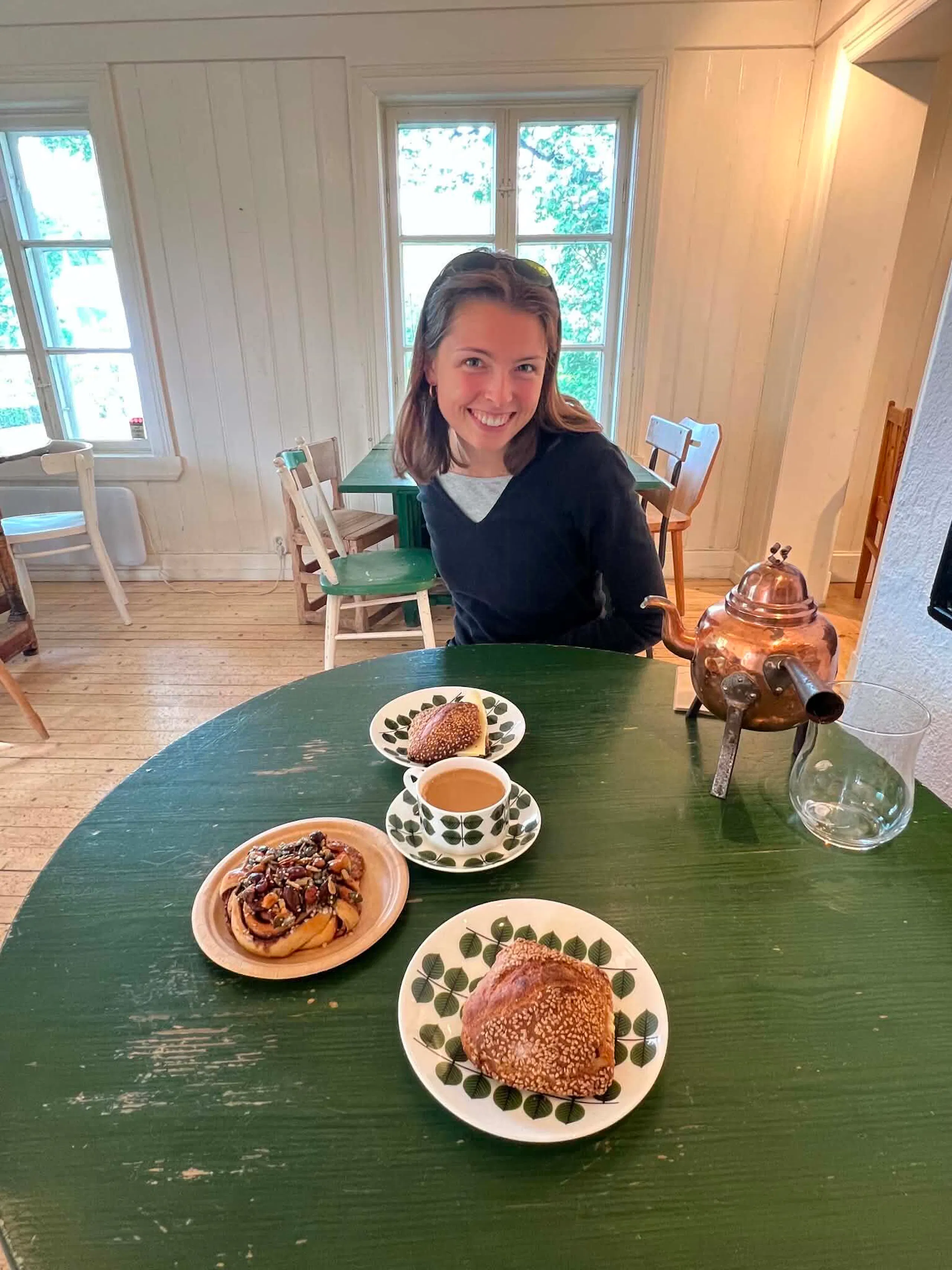 Woman sitting at a green round table with pastries, coffee, and a copper teapot in a bright wood-paneled room with windows and simple chairs