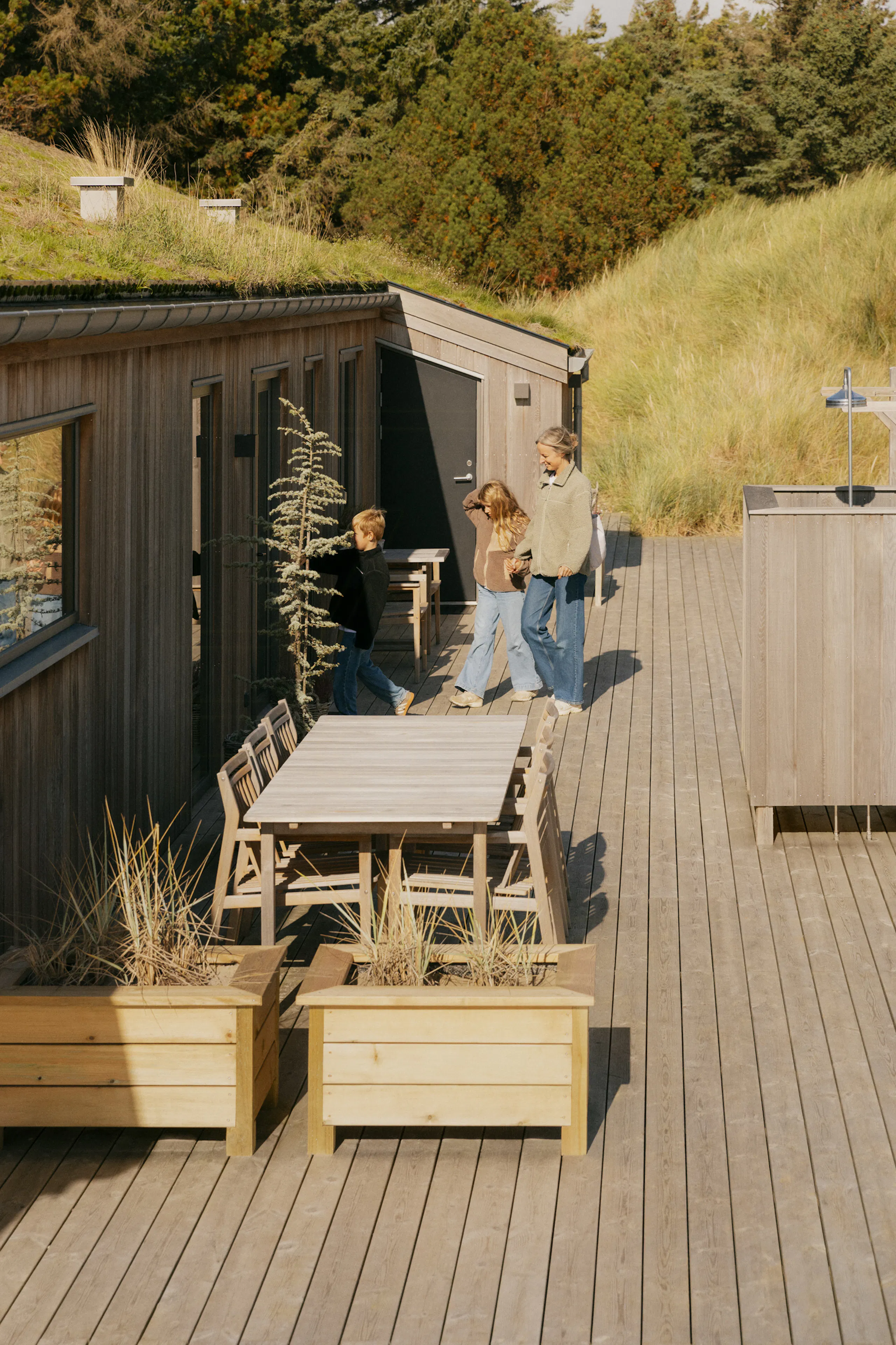 Holzterrasse neben einer niedrigen Hütte mit Grasdach, Esstisch und Stühlen im Freien, Pflanzkästen und drei Personen am Eingang, umgeben von hohen Dünen und Bäumen