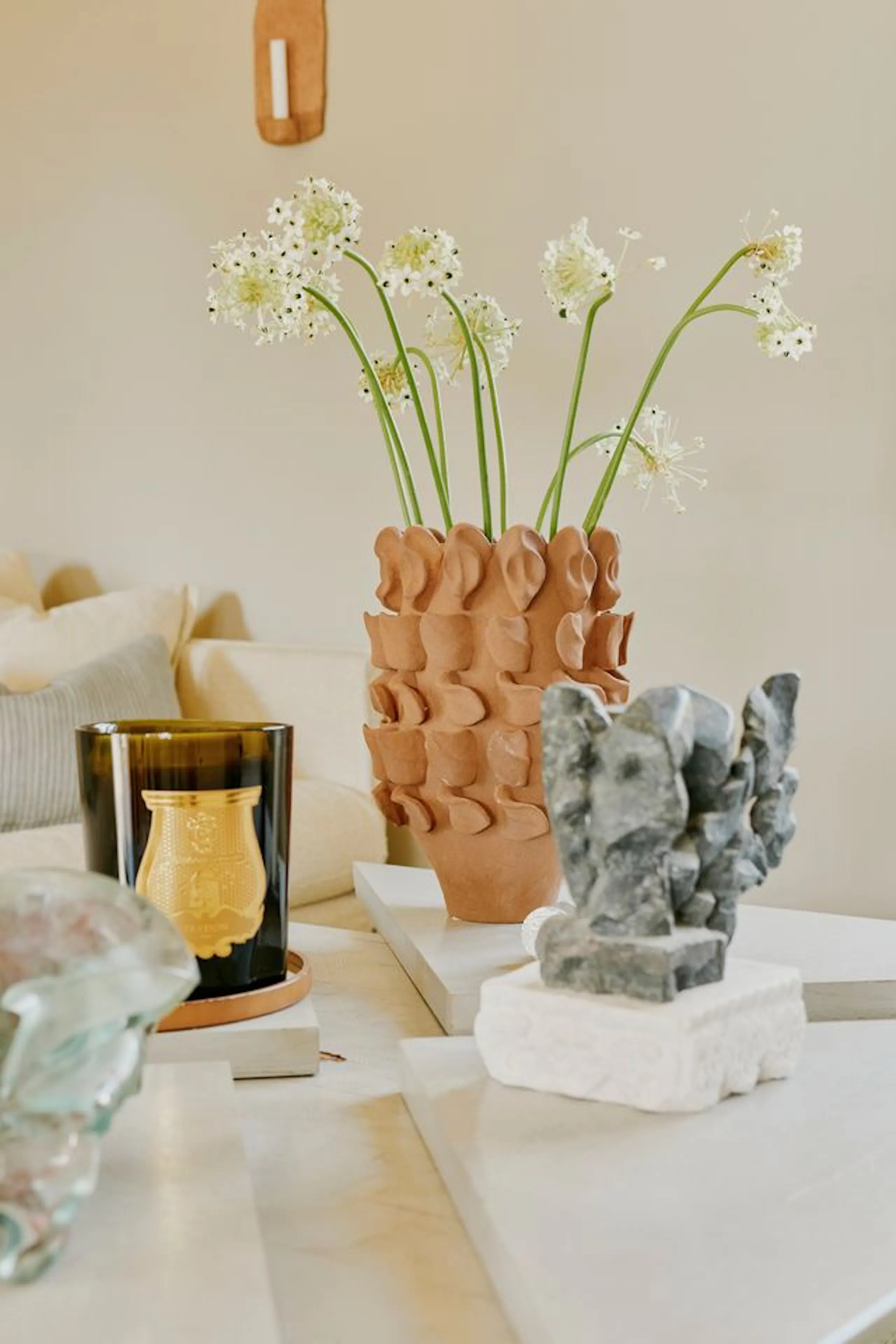 Tabletop arrangement with a terracotta vase of white flowers, a dark glass candle, and small stone sculptures on a light wooden surface