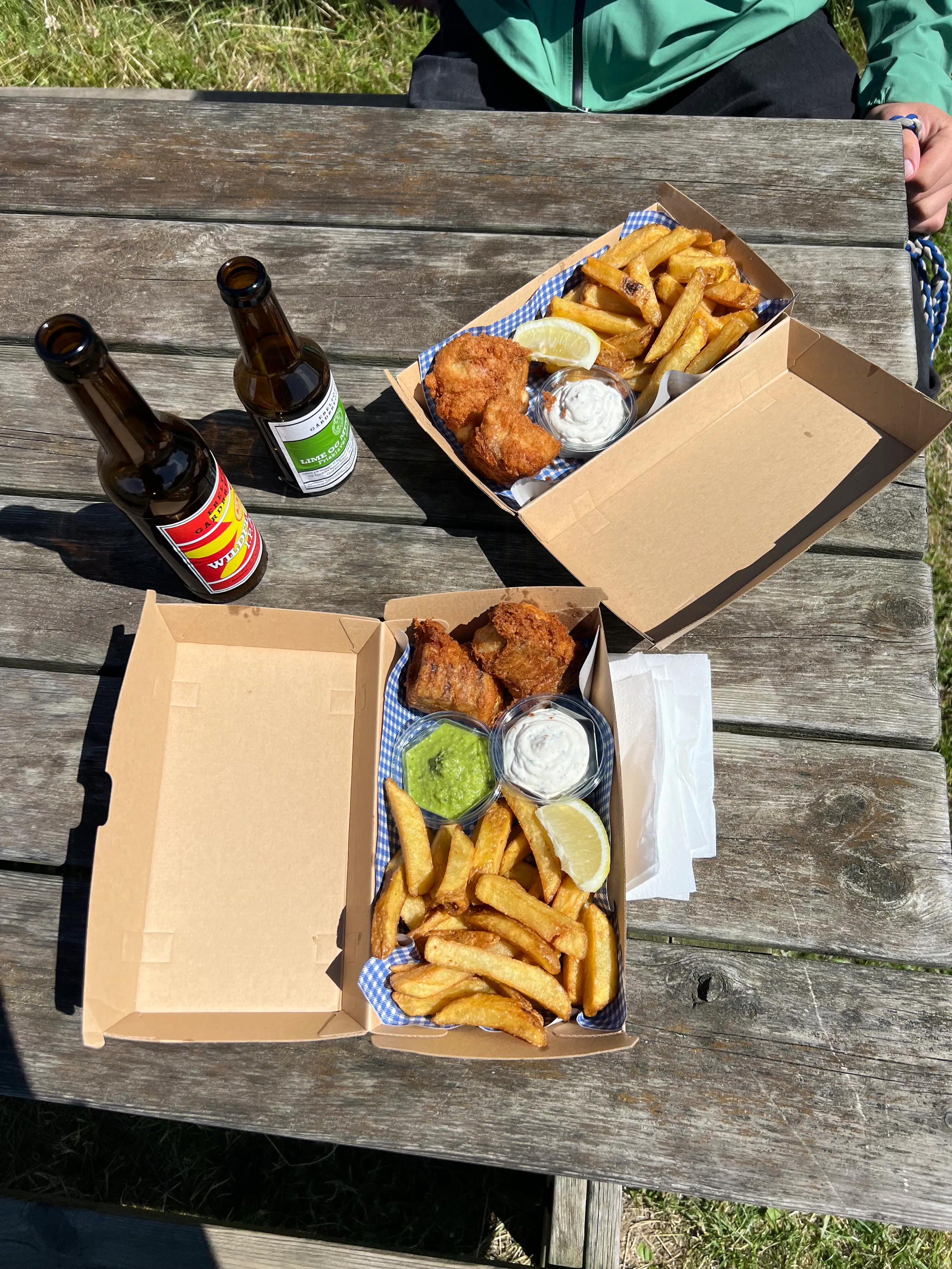 Two takeout boxes with fried chicken, thick-cut fries, lemon wedges, and dipping sauces on a weathered wooden picnic table, with two beer bottles nearby and a person in a green jacket at the top edge