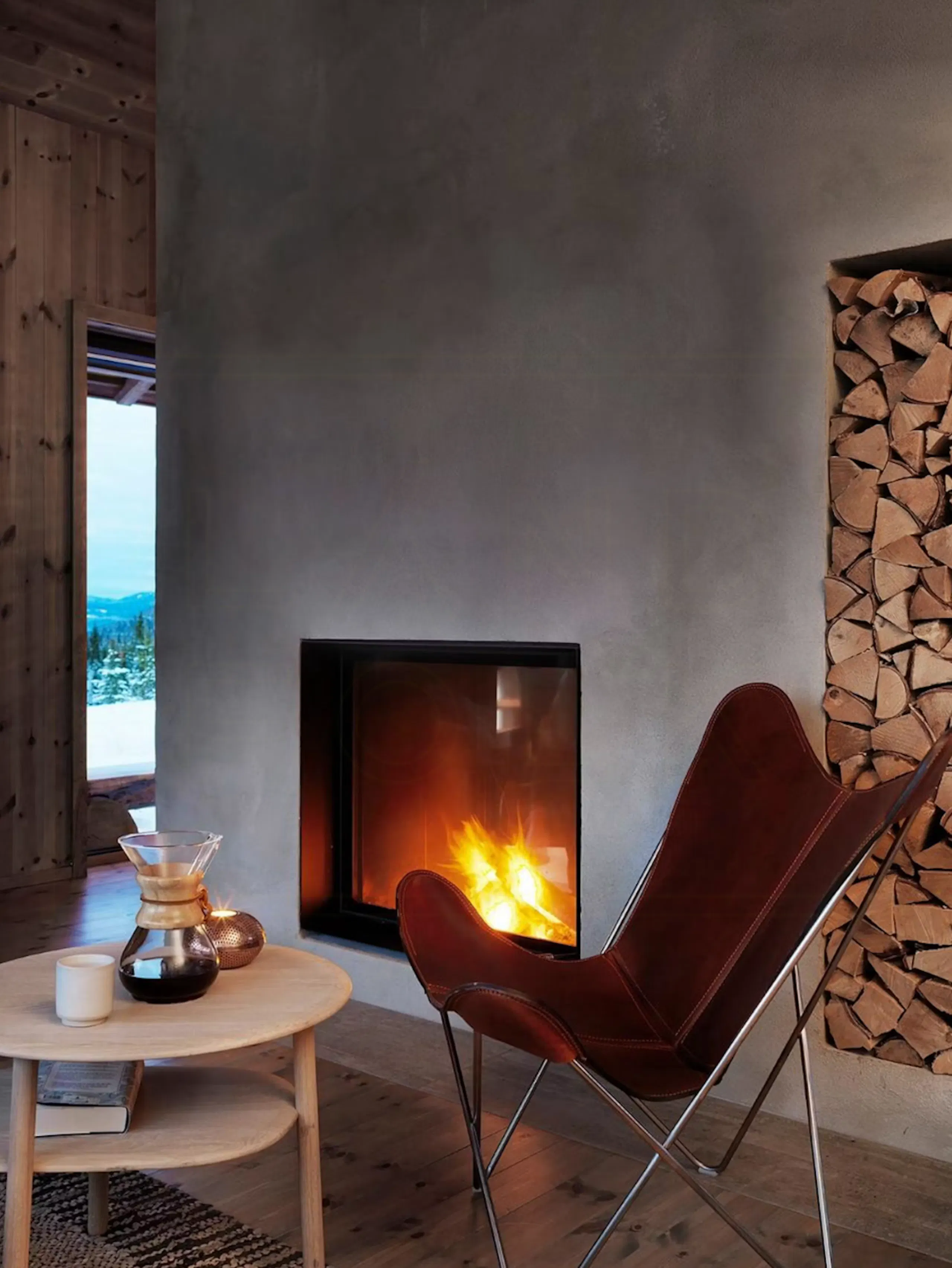 Cozy living room with a lit modern fireplace, leather butterfly chair, round coffee table, stacked firewood niche, and wooden walls with a view outside
