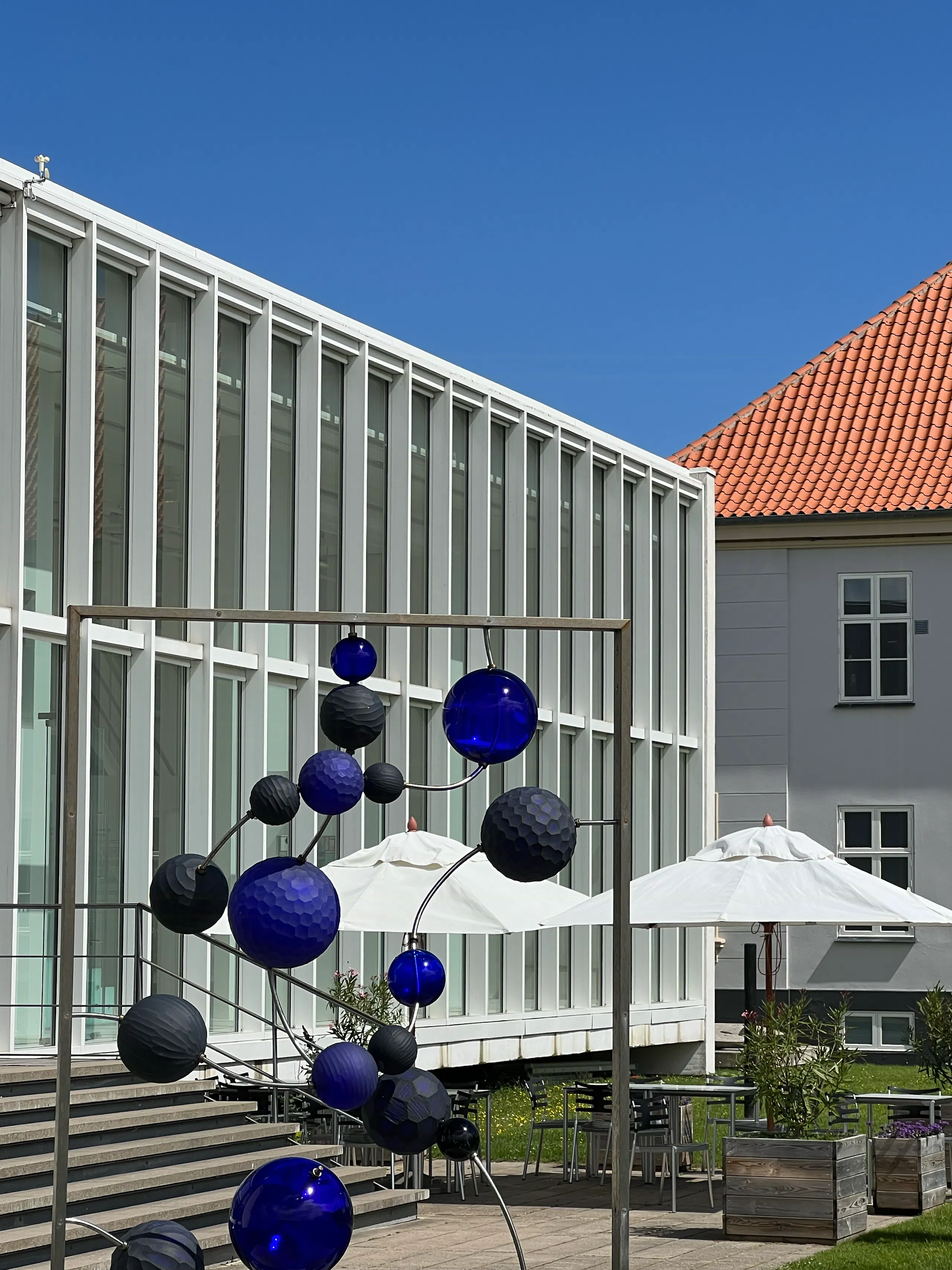 Outdoor terrace with white umbrellas, tables, wooden planters, a modern glass-fronted building, and a metal sculpture with blue and black spheres against a red-tiled roof and clear sky
