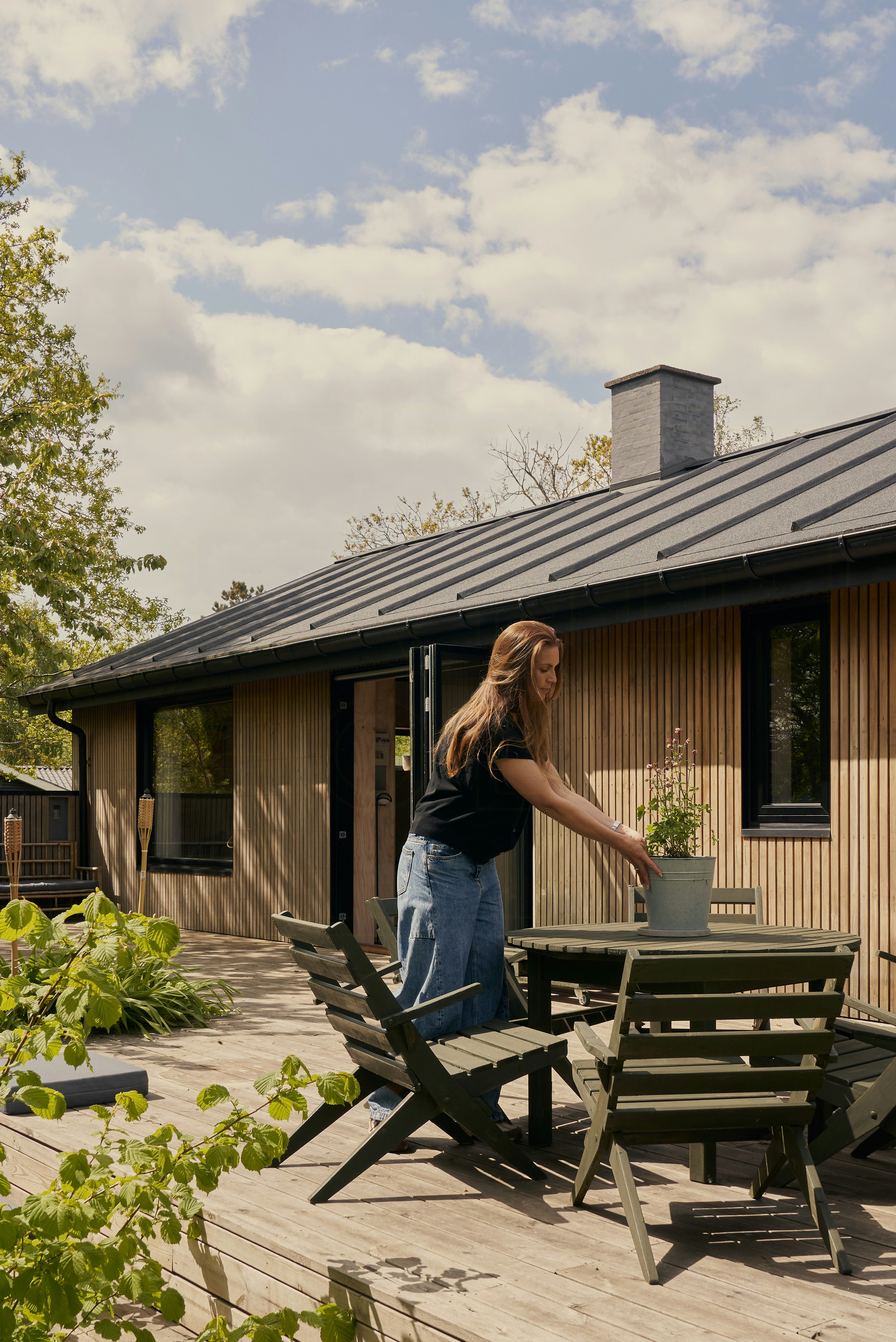 Frau stellt eine Topfpflanze auf einen runden Gartentisch auf einer Holzterrasse neben einem modernen, holzverkleideten Ferienhaus