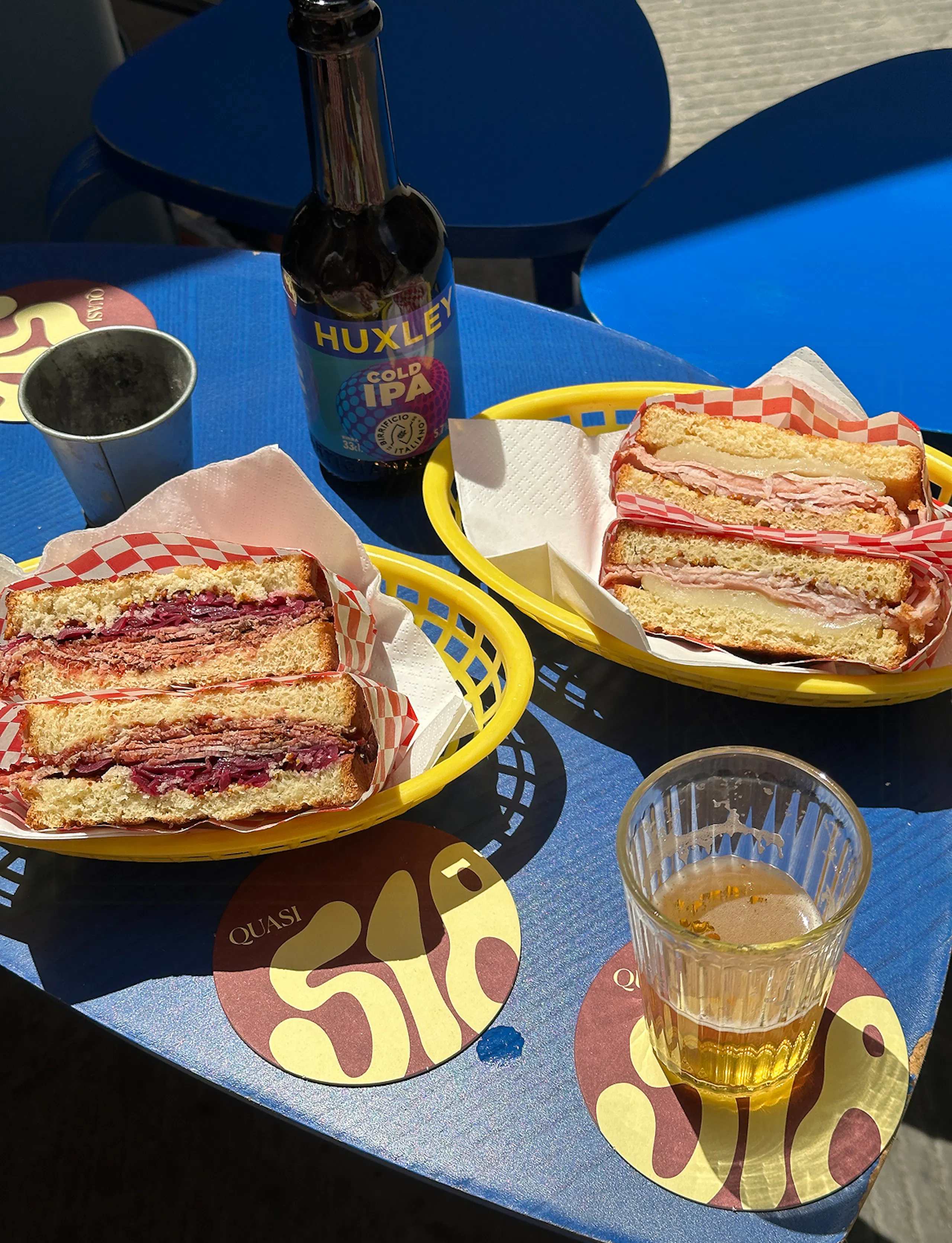 Two sandwiches in yellow baskets on a blue table, with a Huxley Cold IPA bottle, a metal cup, and a half-full glass on coasters outdoors in sunlight