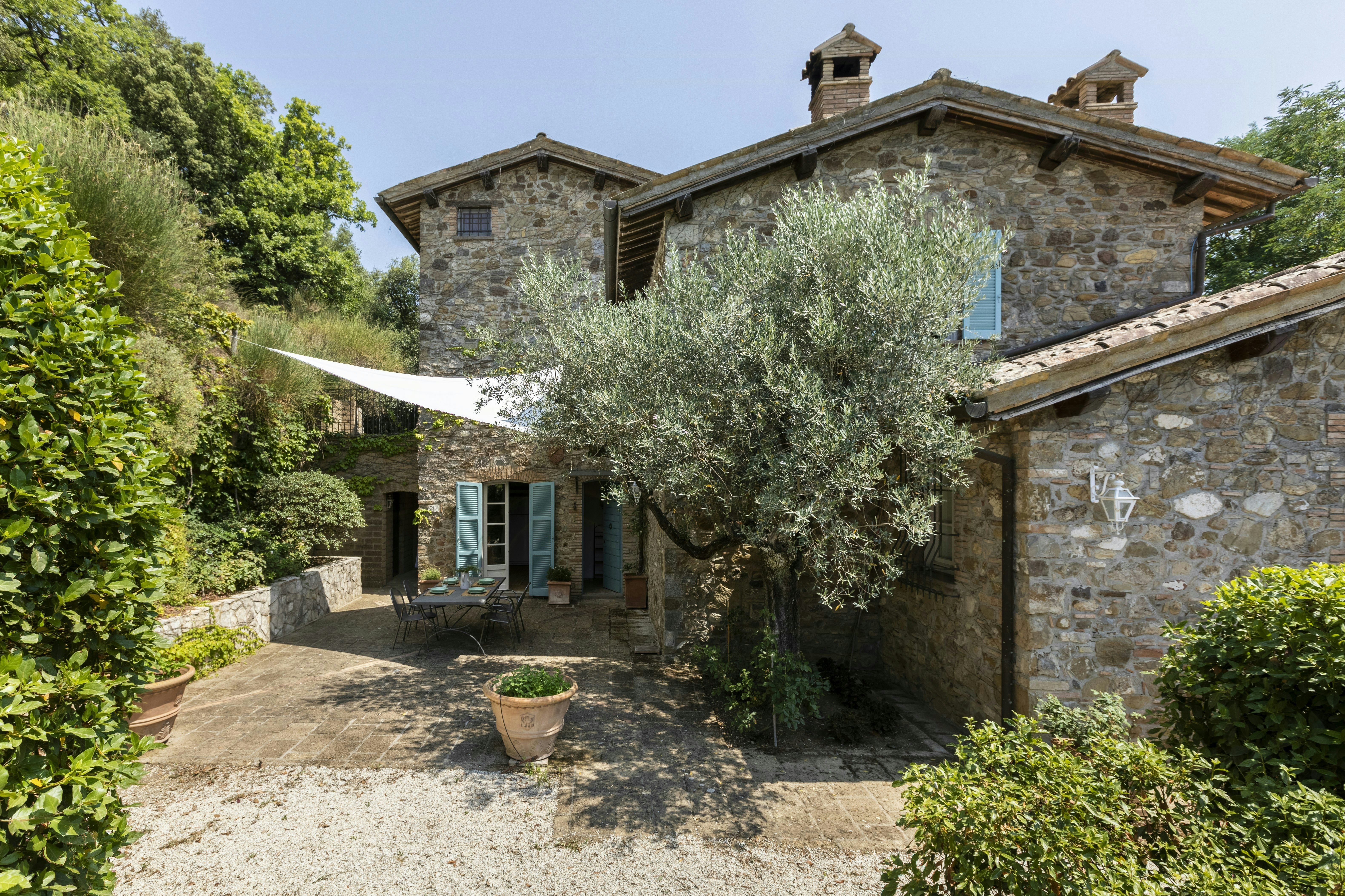 Stone house with blue shutters, small patio with outdoor table and chairs, white shade sail, and an olive tree in front, surrounded by greenery