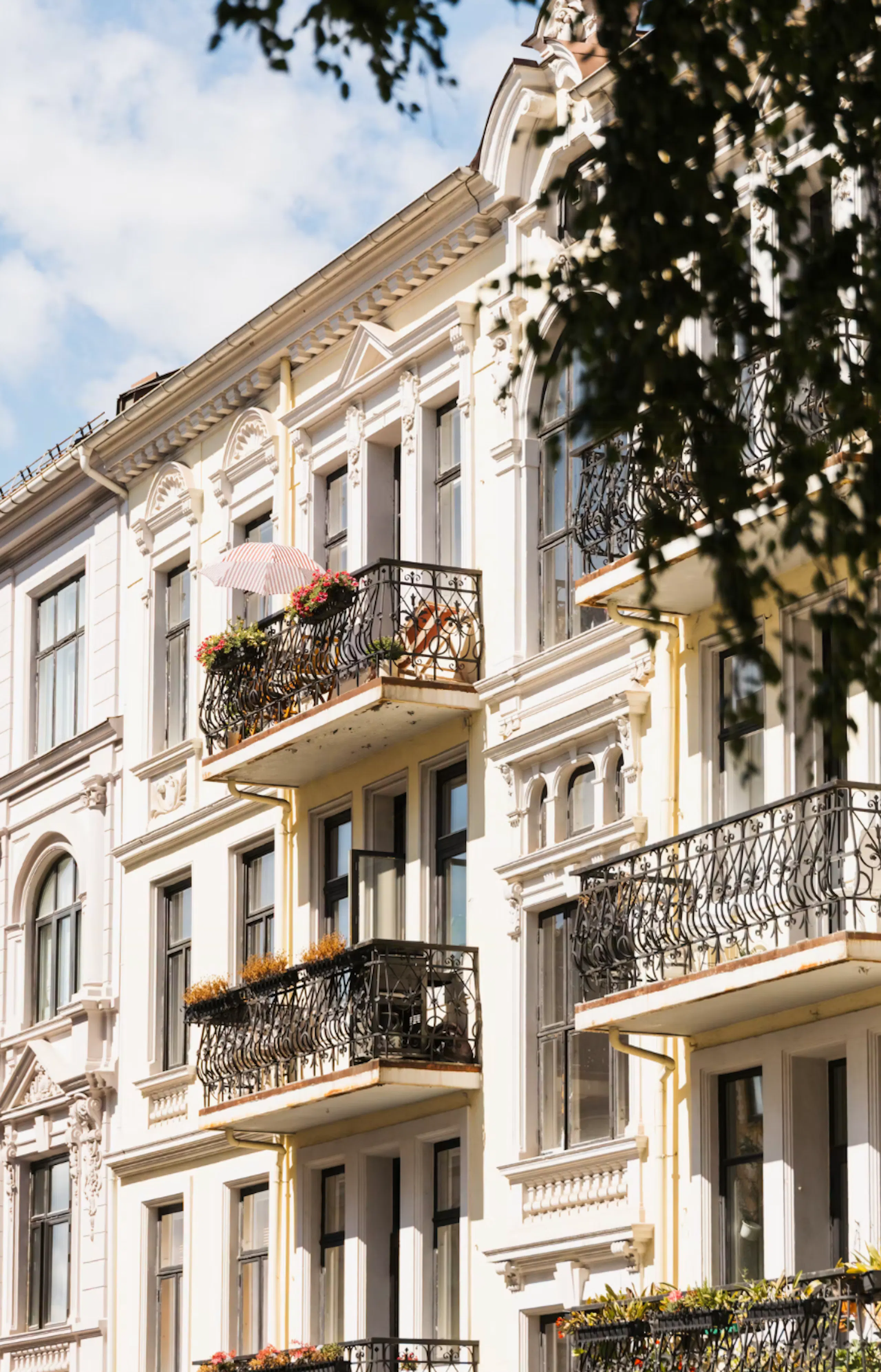 White ornate apartment building with wrought-iron balconies, flower boxes, and a pink-striped umbrella, framed by tree branches against a blue sky