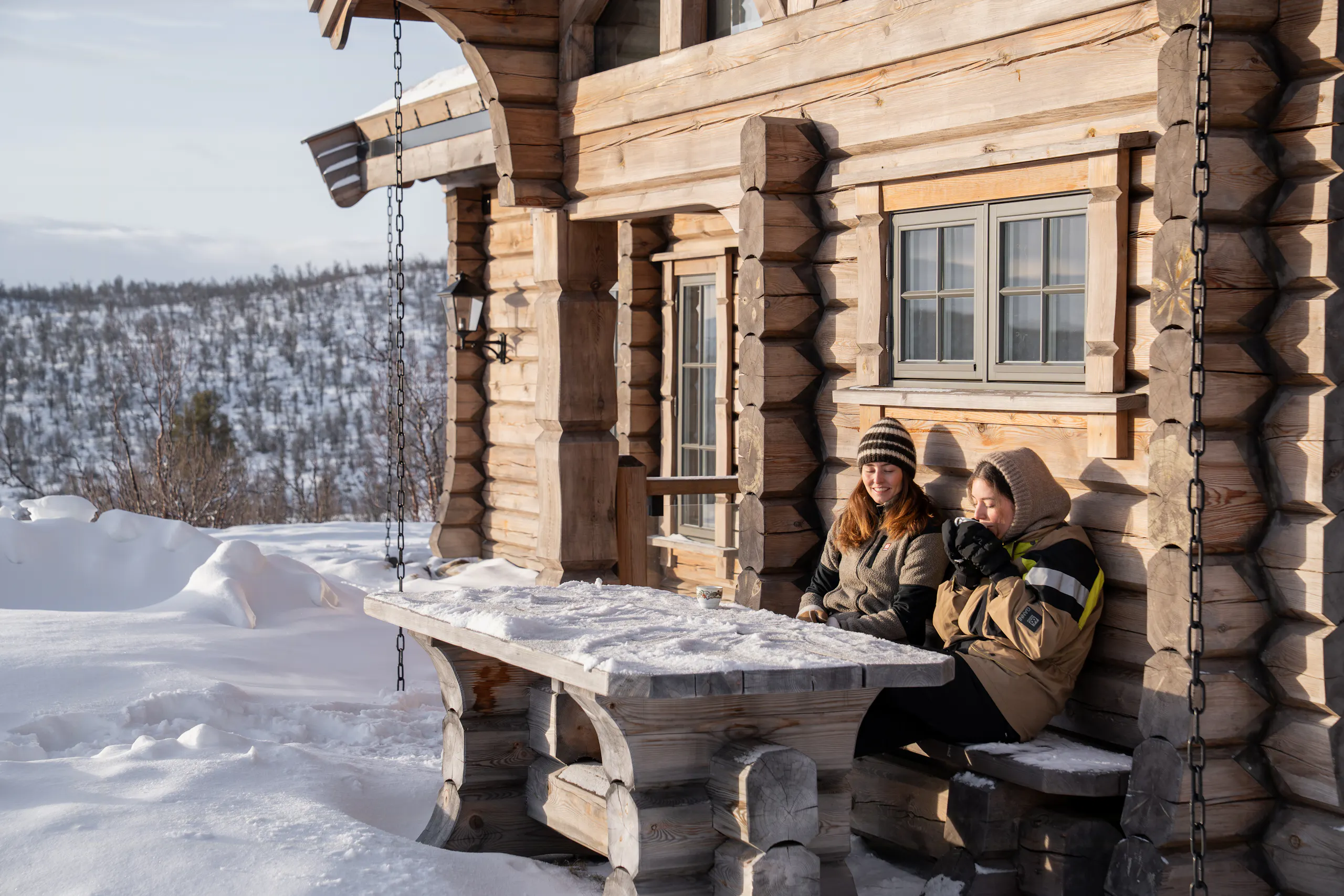 Two people sitting on a snow-covered wooden bench by a log cabin porch, with a snowy forest landscape in the background