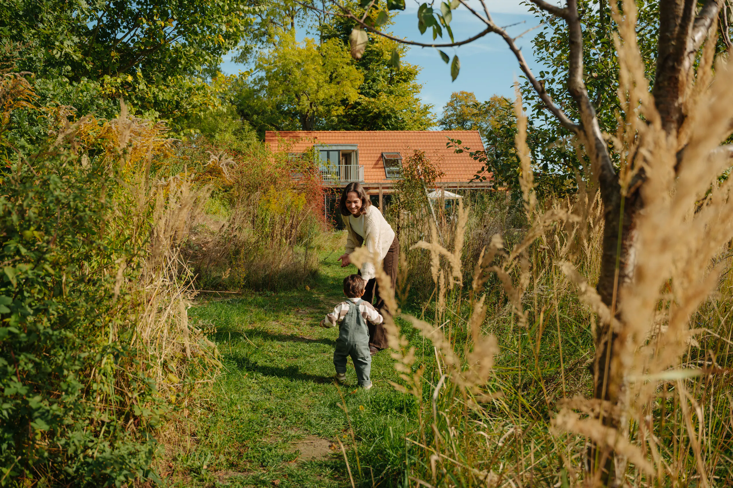 Frau mit einem kleinen Kind auf einem grasbewachsenen Gartenweg, hohe Wildgräser und Bäume im Vordergrund und ein Haus mit rotem Dach im Hintergrund