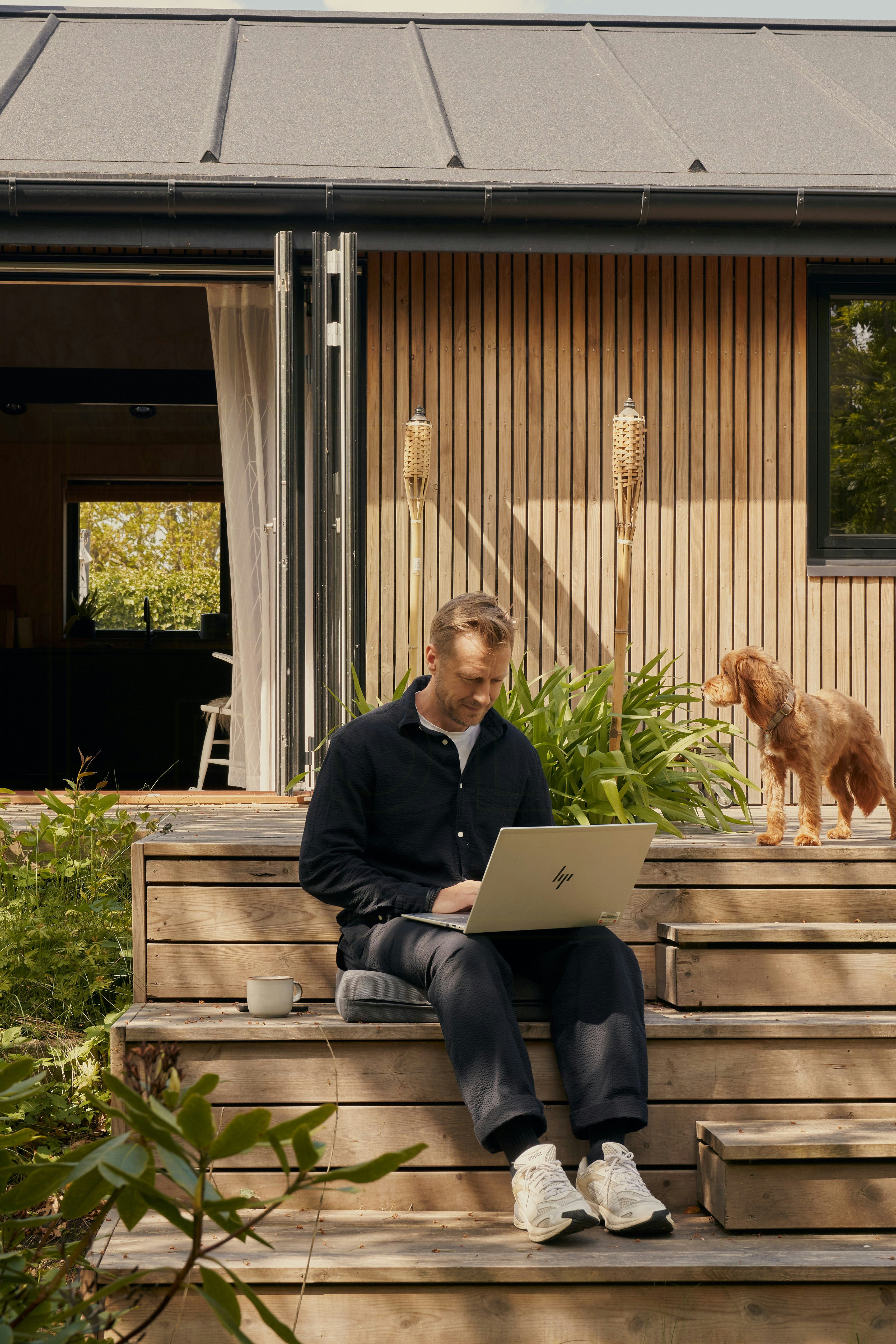 Mann sitzt auf Holzstufen und arbeitet an einem Laptop neben einem kleinen braunen Hund vor einem holzverkleideten Ferienhaus mit offenen Türen