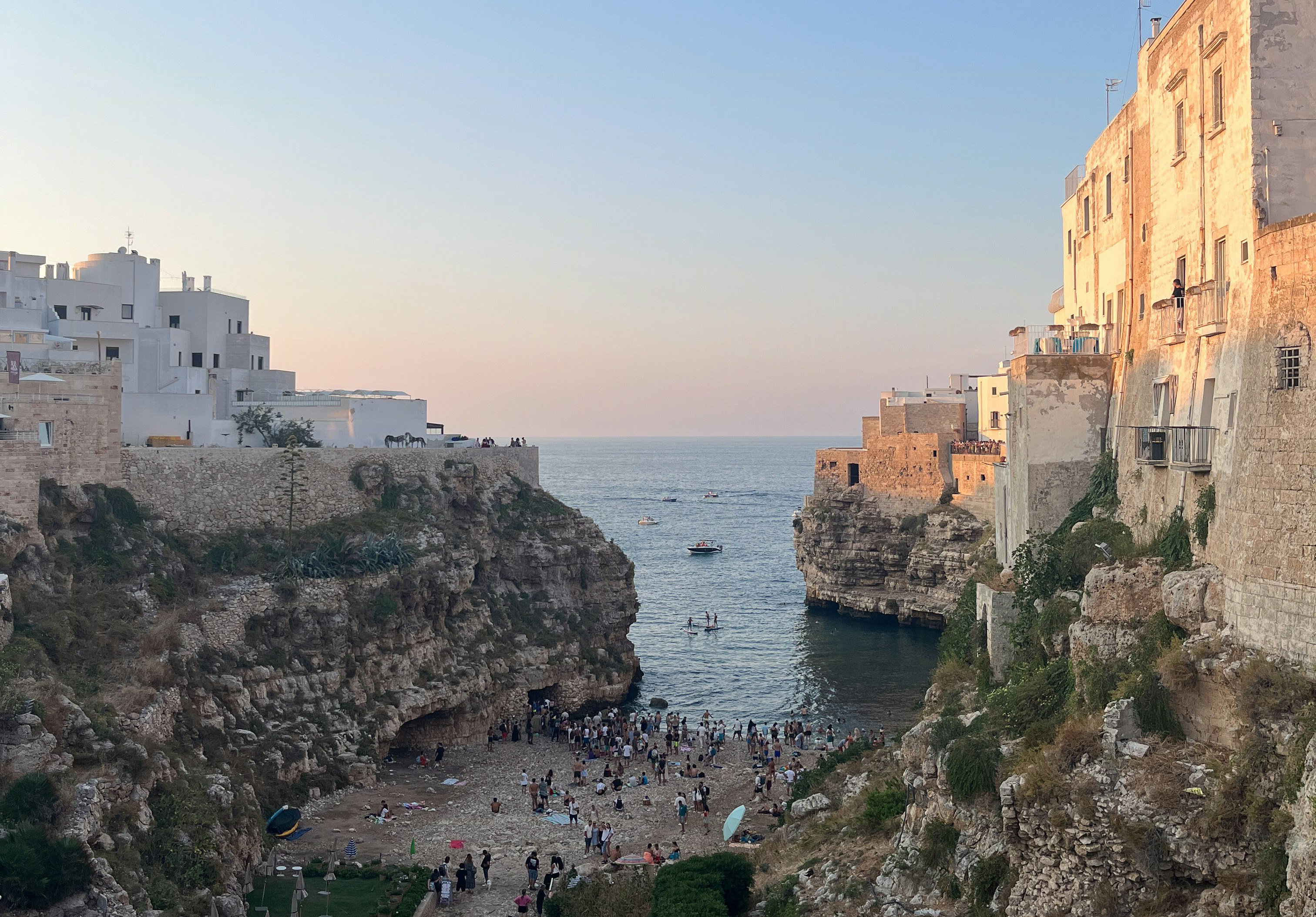 Rocky coastal cove with small beach and swimmers, high stone buildings on cliffs at both sides, and boats on calm sea at sunset
