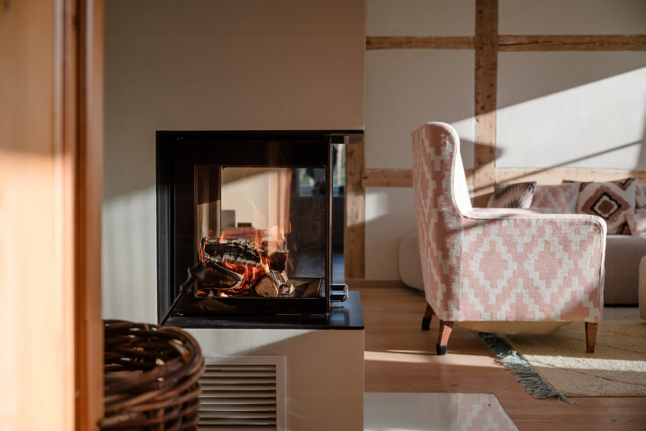 Modern fireplace with burning logs next to a patterned armchair and sofa in a sunlit living room with wooden floors and exposed beam walls