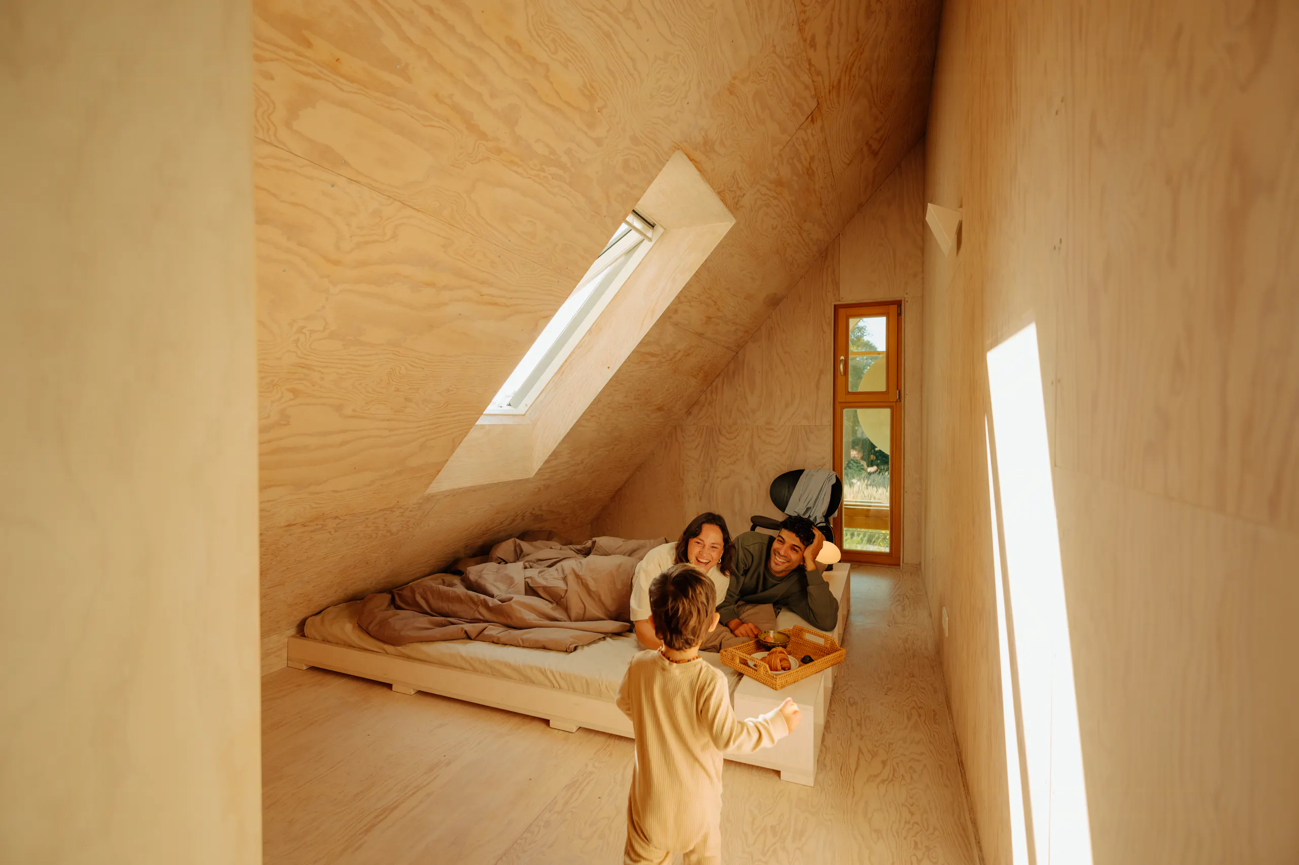 Mit Sperrholz verkleidetes Schlafzimmer im Dachgeschoss mit schrägen Wänden, zwei Erwachsenen im Bett, einem daneben stehenden Kind, einem Dachfenster und schmalen senkrechten Fenstern, die Sonnenlicht hereinlassen