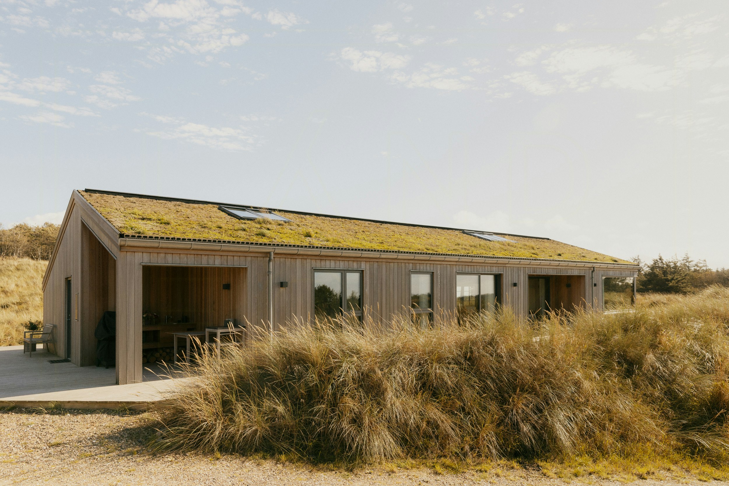 Einstöckiges Holzhaus mit moosbewachsenem Schrägdach, großen Fenstern und überdachter Terrasse, umgeben von hohem Strandgras unter einem hellen Himmel