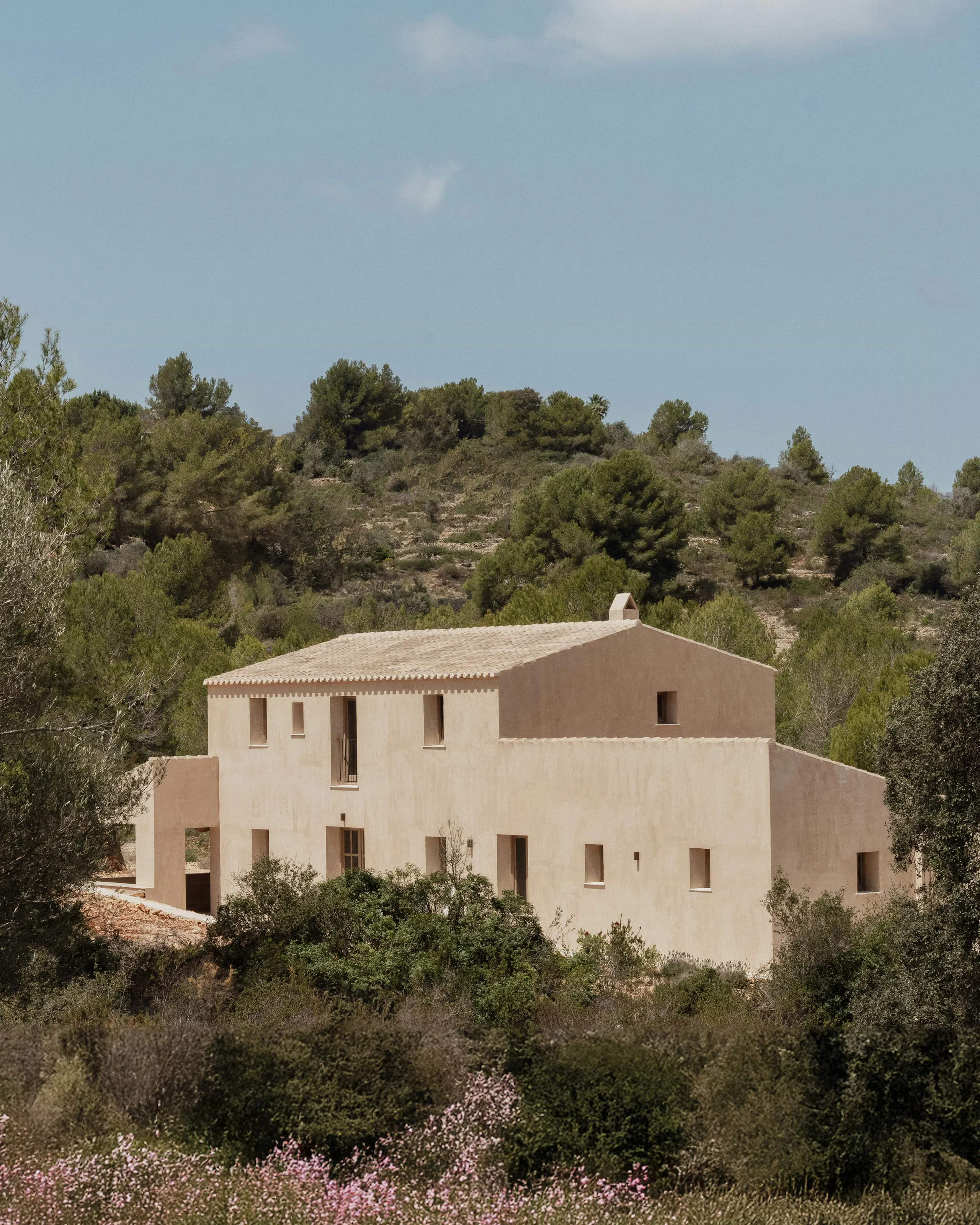 Two-story beige house with a tiled roof and small windows surrounded by trees and shrubs on a hillside under a clear sky