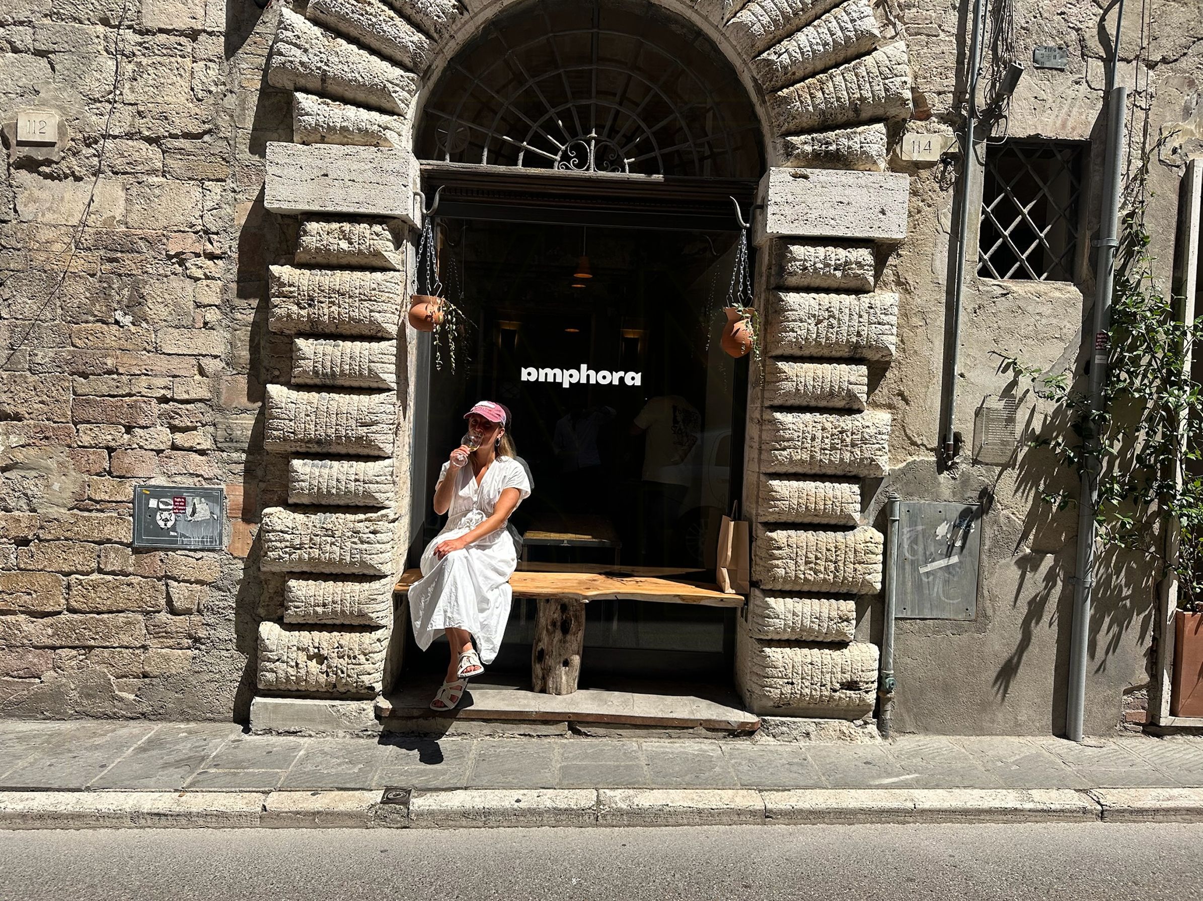 Woman in a white dress and pink cap sitting in a stone doorway with a wooden bench, in front of a dark storefront with the word “amphora” visible