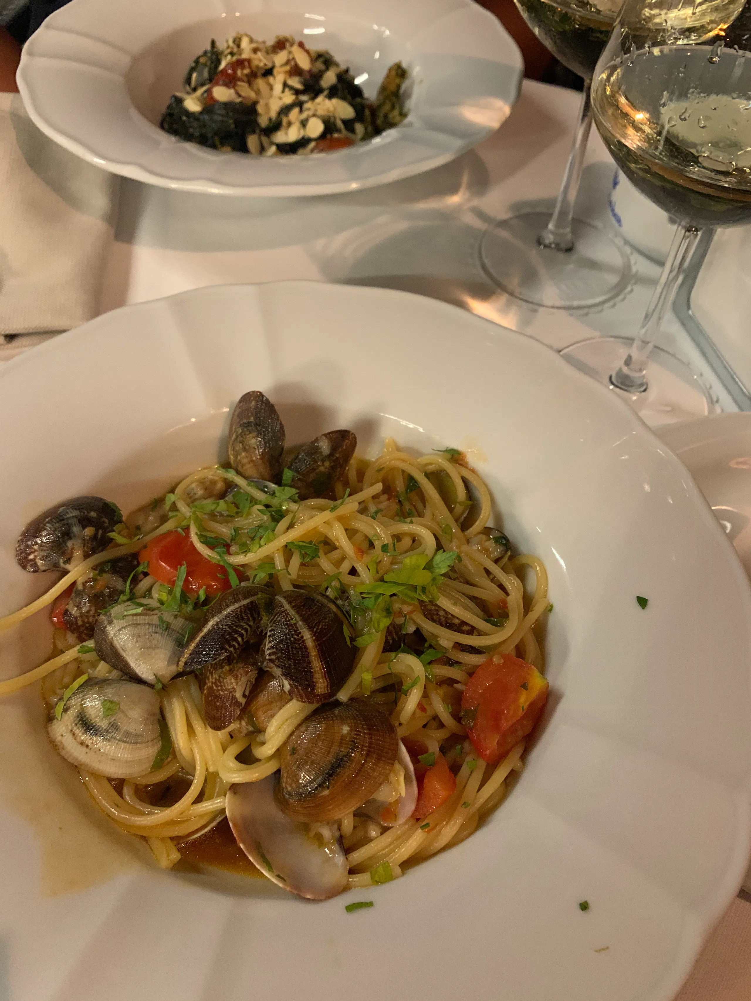 Plate of spaghetti with clams and cherry tomatoes on a white tablecloth, with a second pasta dish and two wine glasses in the background