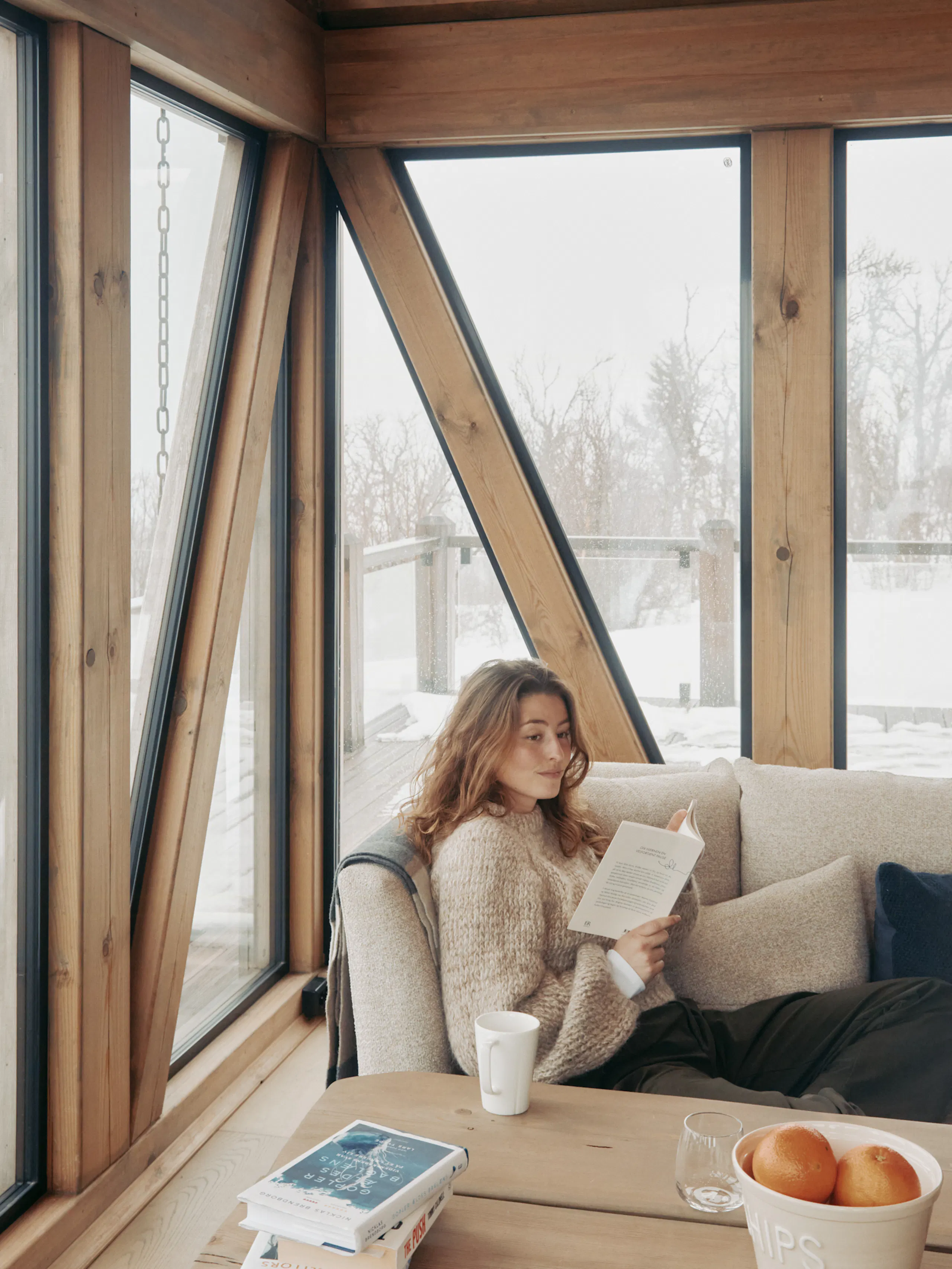 Woman reading on a beige sofa in a wood-framed room with large windows, with snow visible outside and a table with books, mug, glass, and bowl of oranges in front