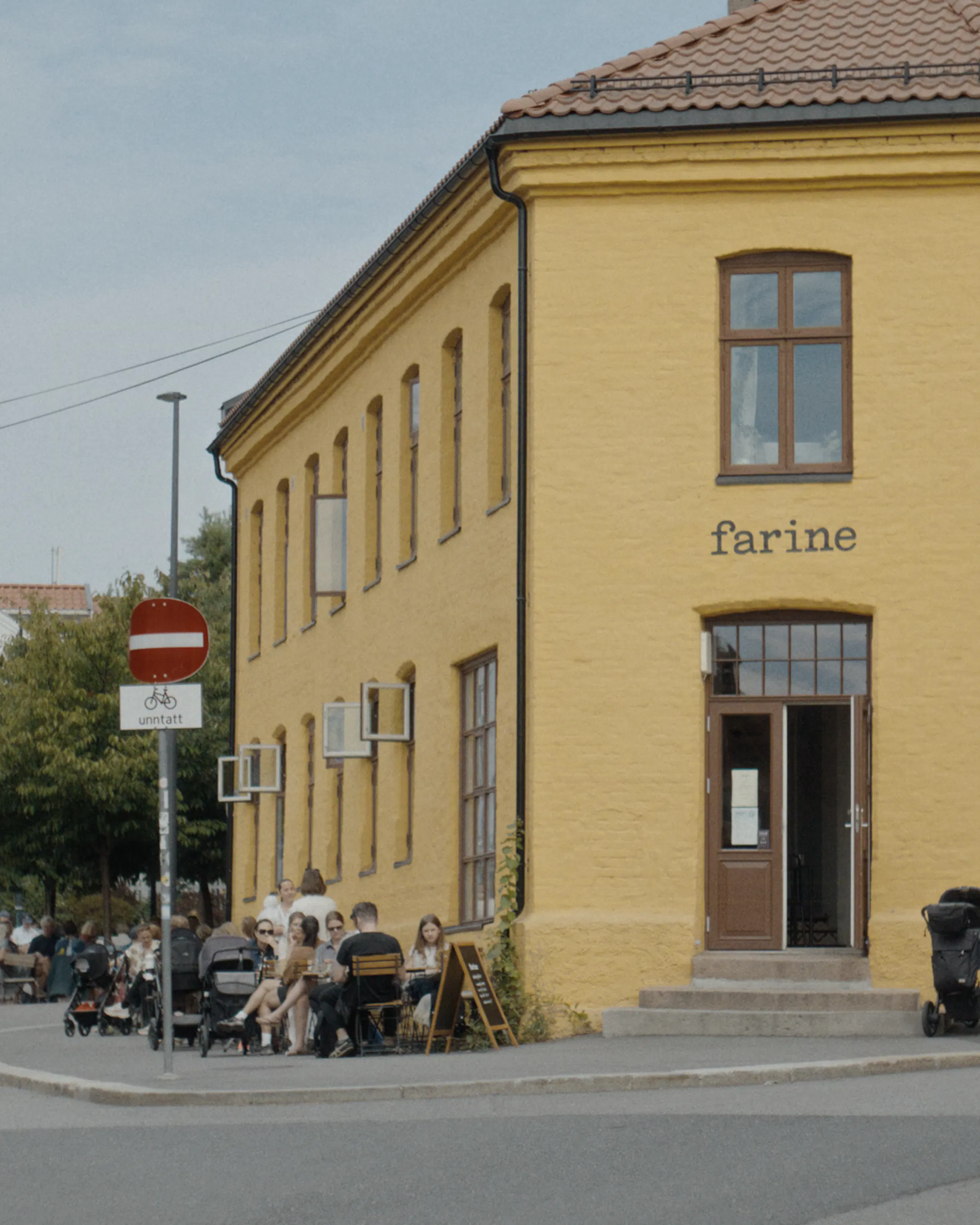 Yellow corner building with brown-framed windows, tiled roof, and the word “farine” above the entrance, with people seated at outdoor café tables beside the sidewalk and street signs nearby