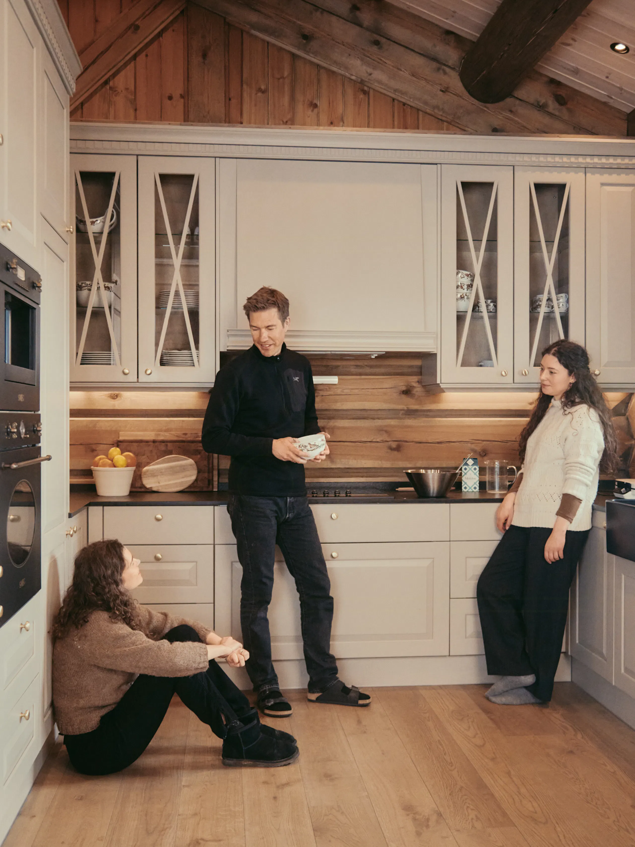 Three people standing and sitting in a wooden kitchen with light cabinets, glass-front cupboards, a built-in oven, and a wood-paneled ceiling