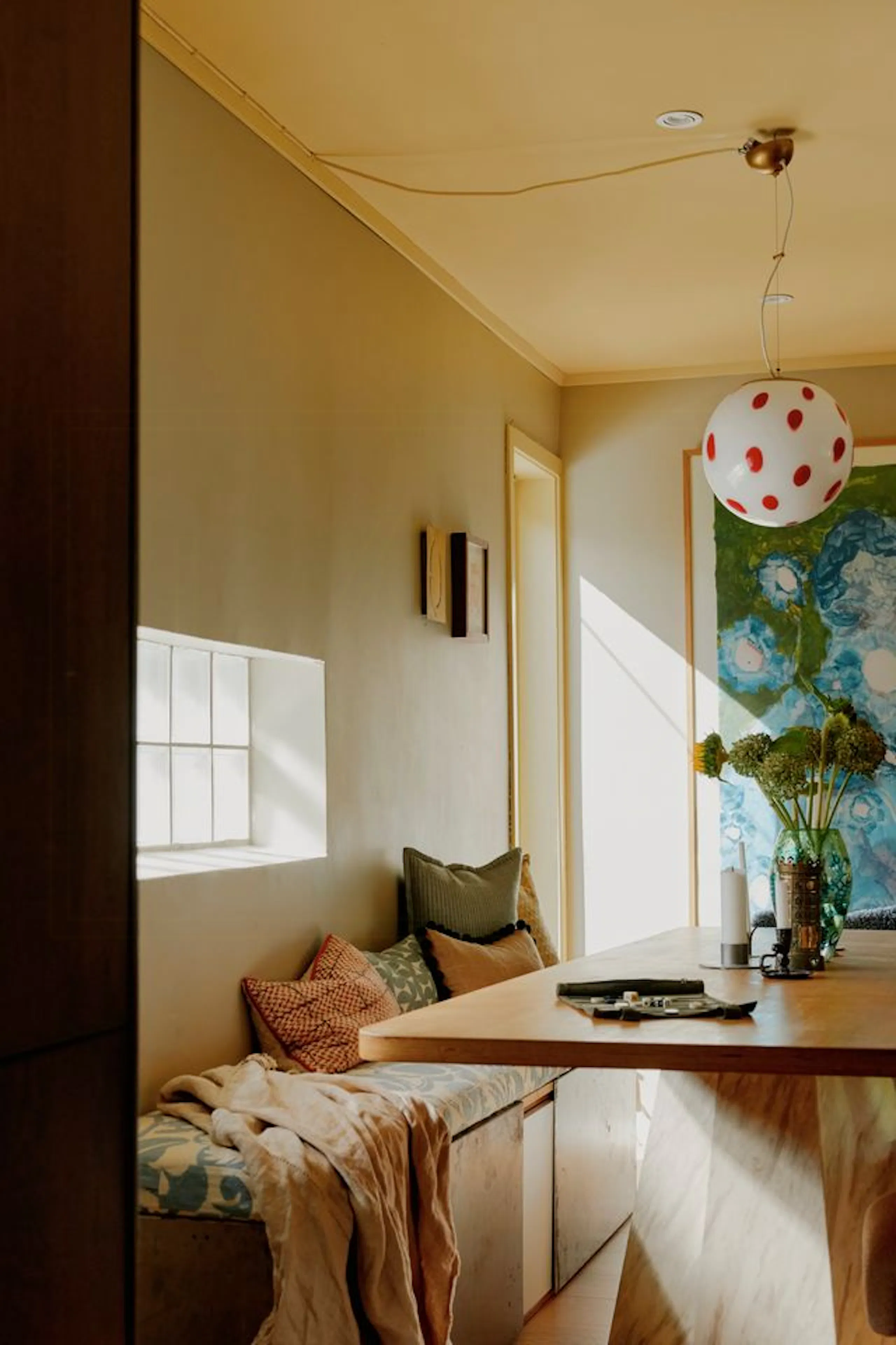 Cozy dining nook with a wooden table, built-in bench seating with cushions and blankets, a round pendant light with red polka dots, and a large blue floral artwork on the wall