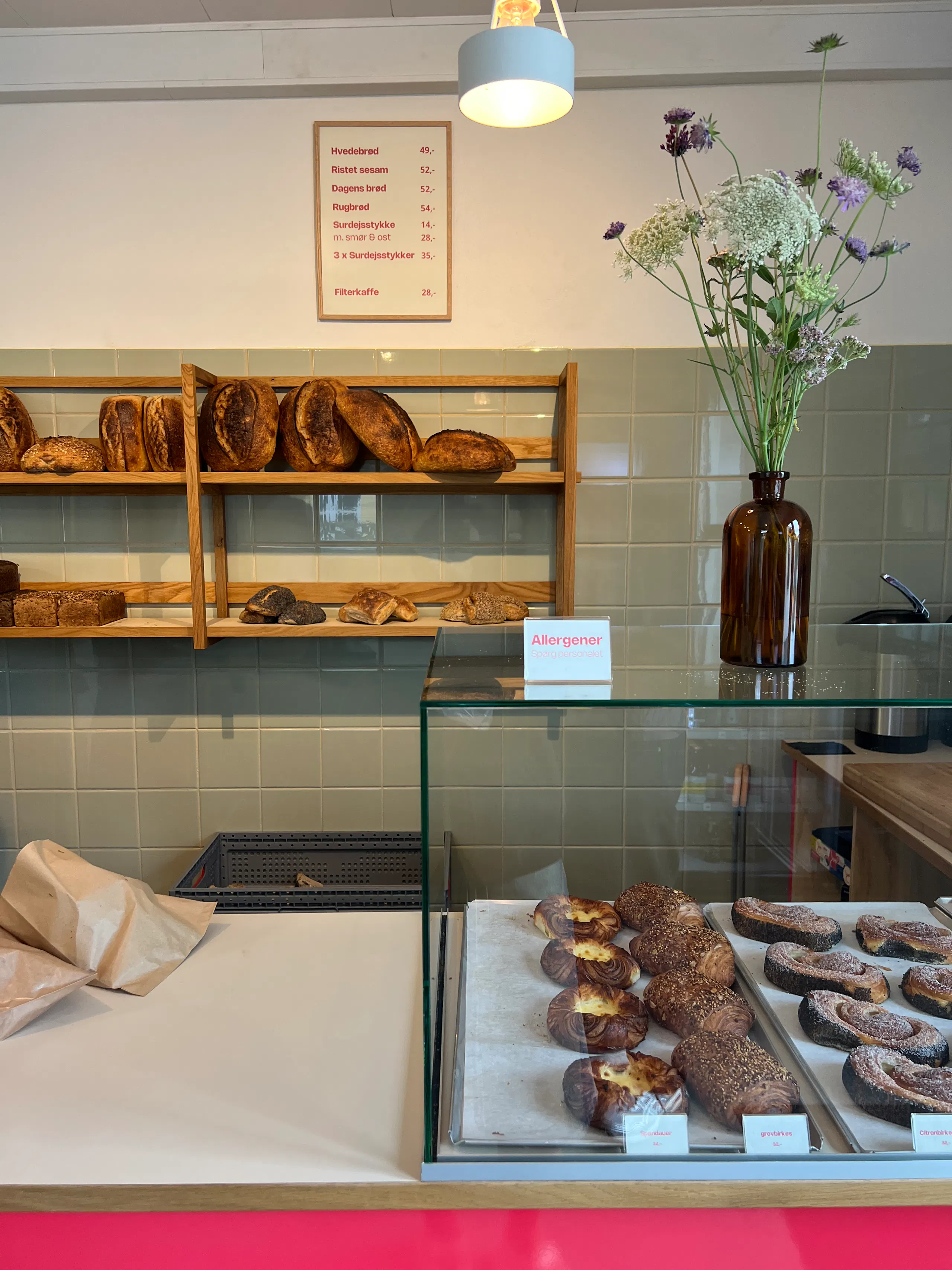 Bakery counter with glass display case of pastries, shelves of loaves behind tiled wall, and a vase of flowers on the counter