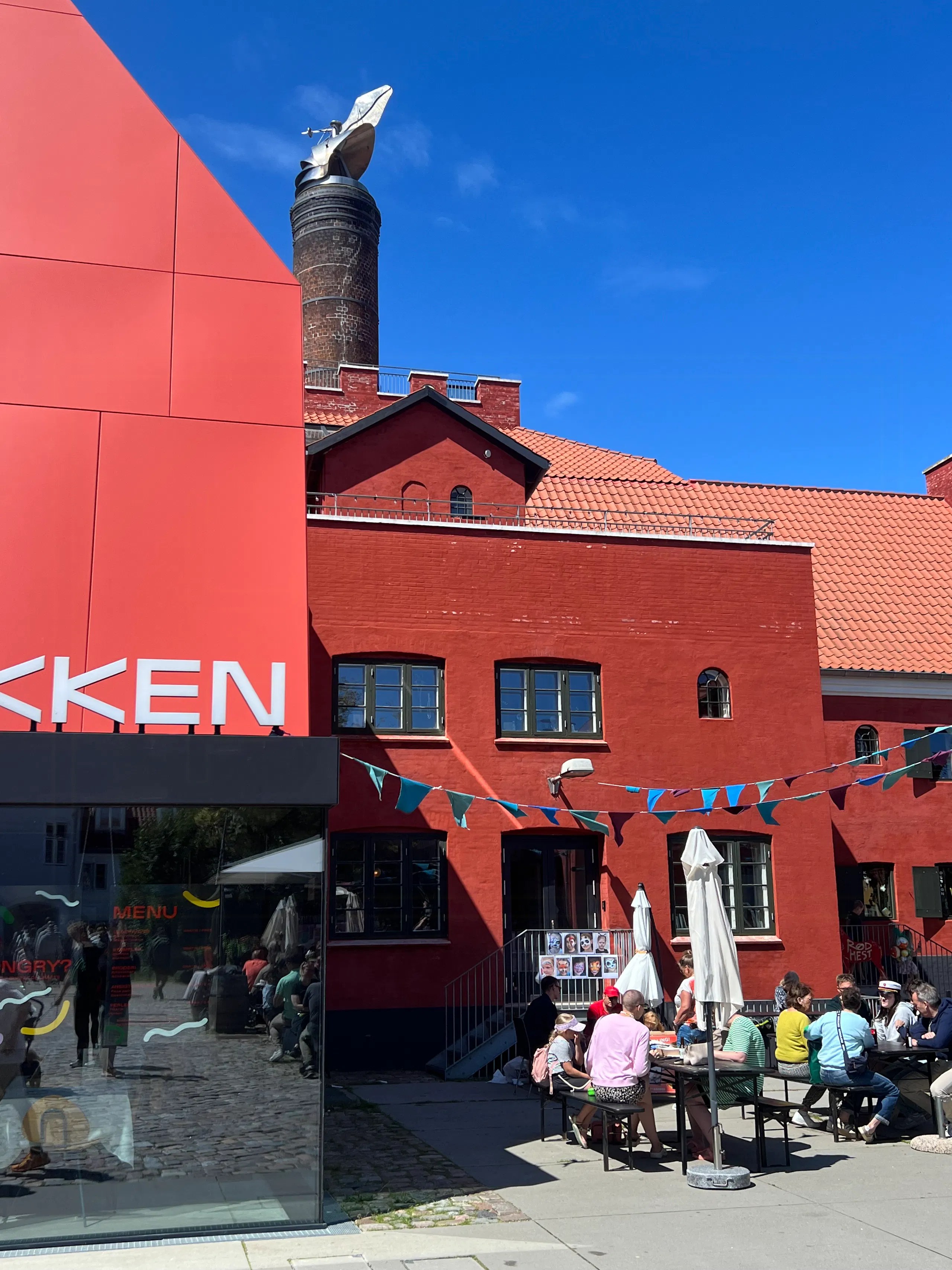 Red brick building with tiled roof and chimney, red modern facade at left, and people sitting at outdoor café tables under umbrellas in a sunny courtyard