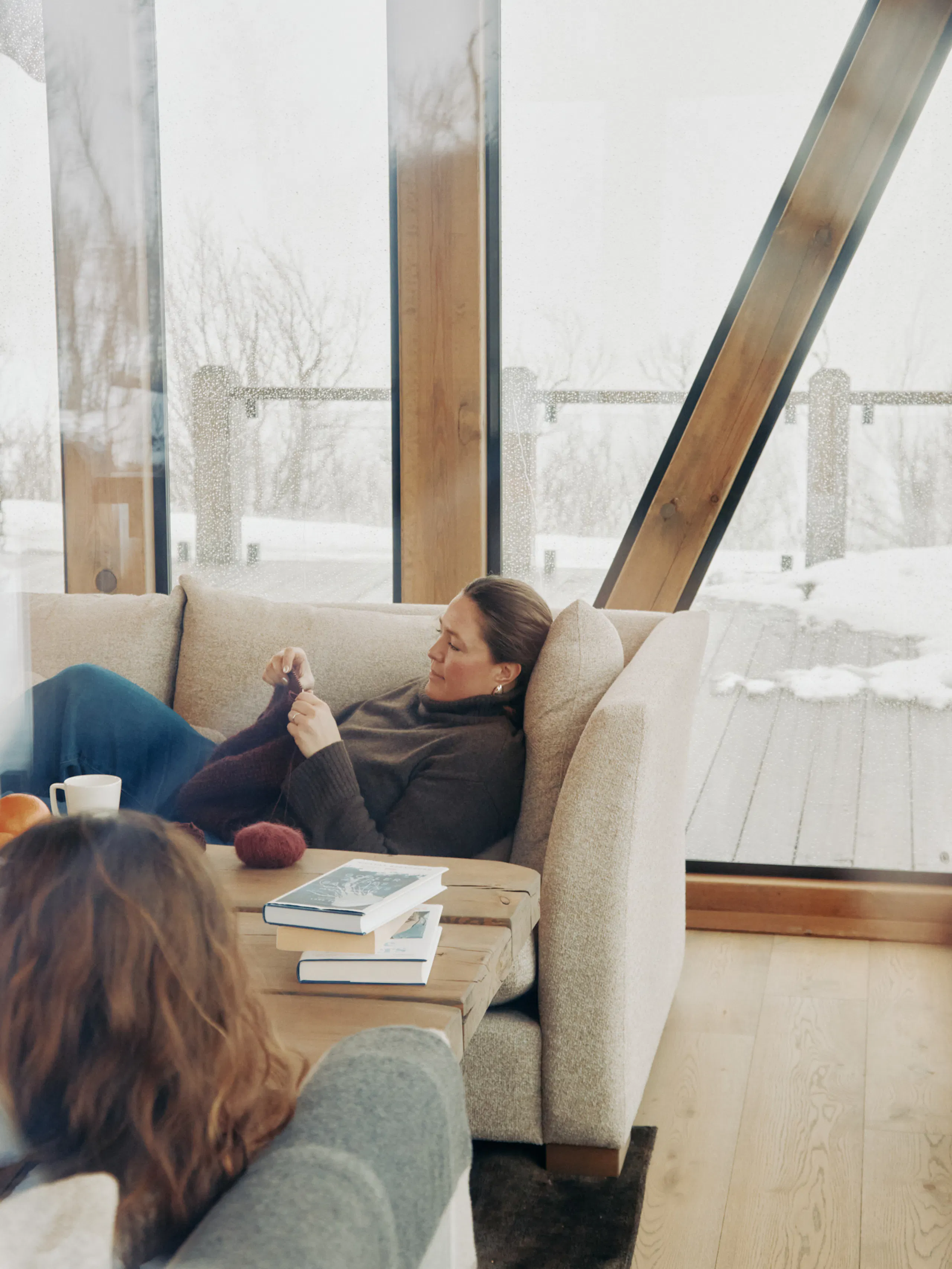 Two people sitting on beige sofas in a bright living room with floor-to-ceiling windows, wooden beams, a wooden coffee table with books and mugs, and snow visible outside