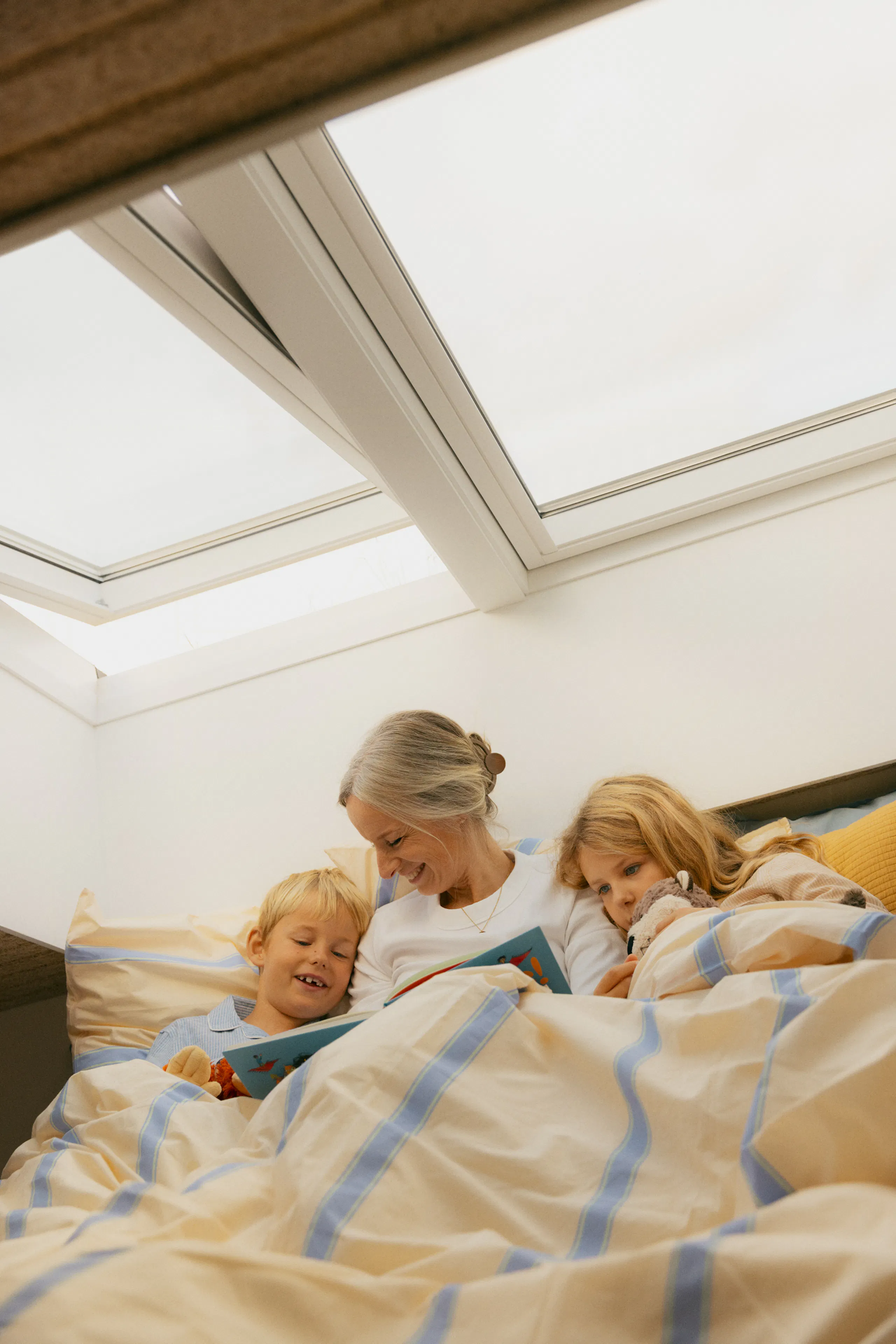 Ältere Frau liest mit zwei Kindern im Bett unter Dachfenstern ein Buch, zugedeckt mit gestreifter Bettdecke und Kissen